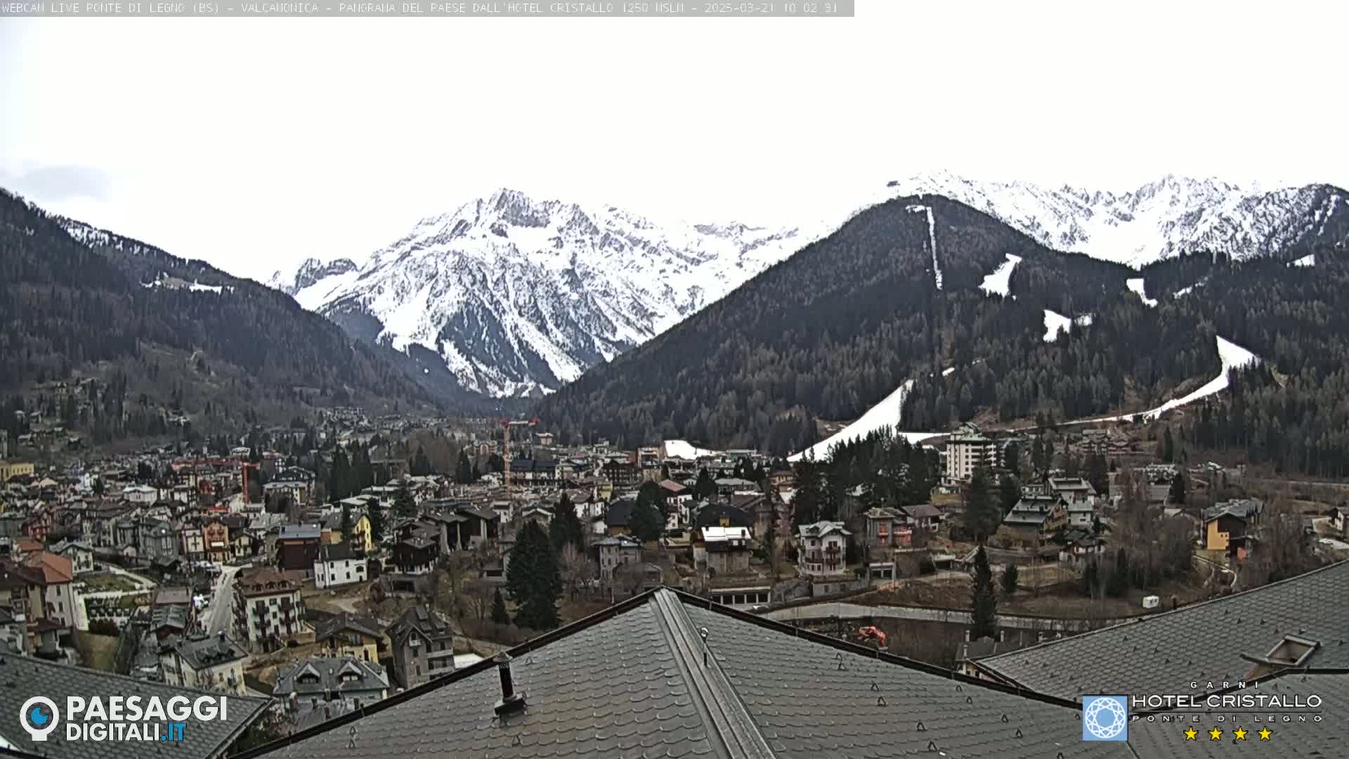 A snow-covered mountain range overlooks a town of closely clustered buildings, under a cloudy sky.