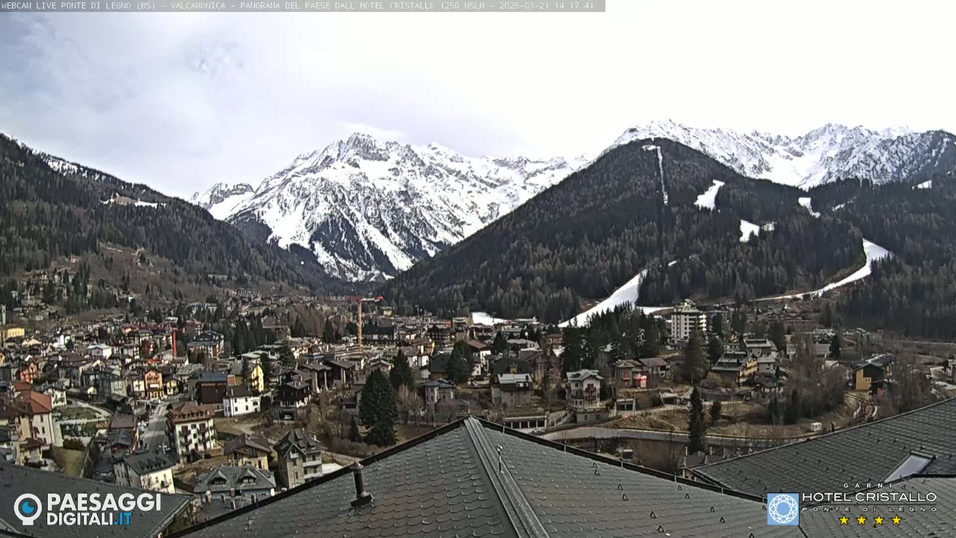 A mountain town nestled in a valley, with snow-capped mountains in the background under a mostly cloudy sky.