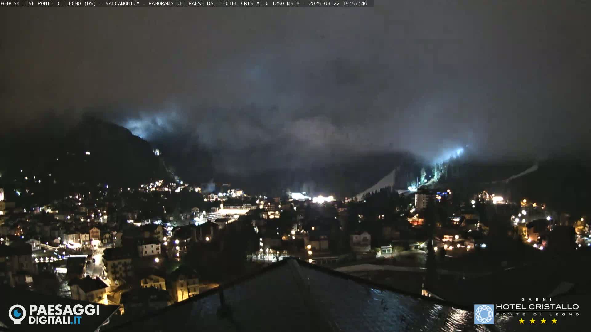 A nighttime view of a town nestled in a valley, partially shrouded in fog or low clouds, with hills illuminated by lights.