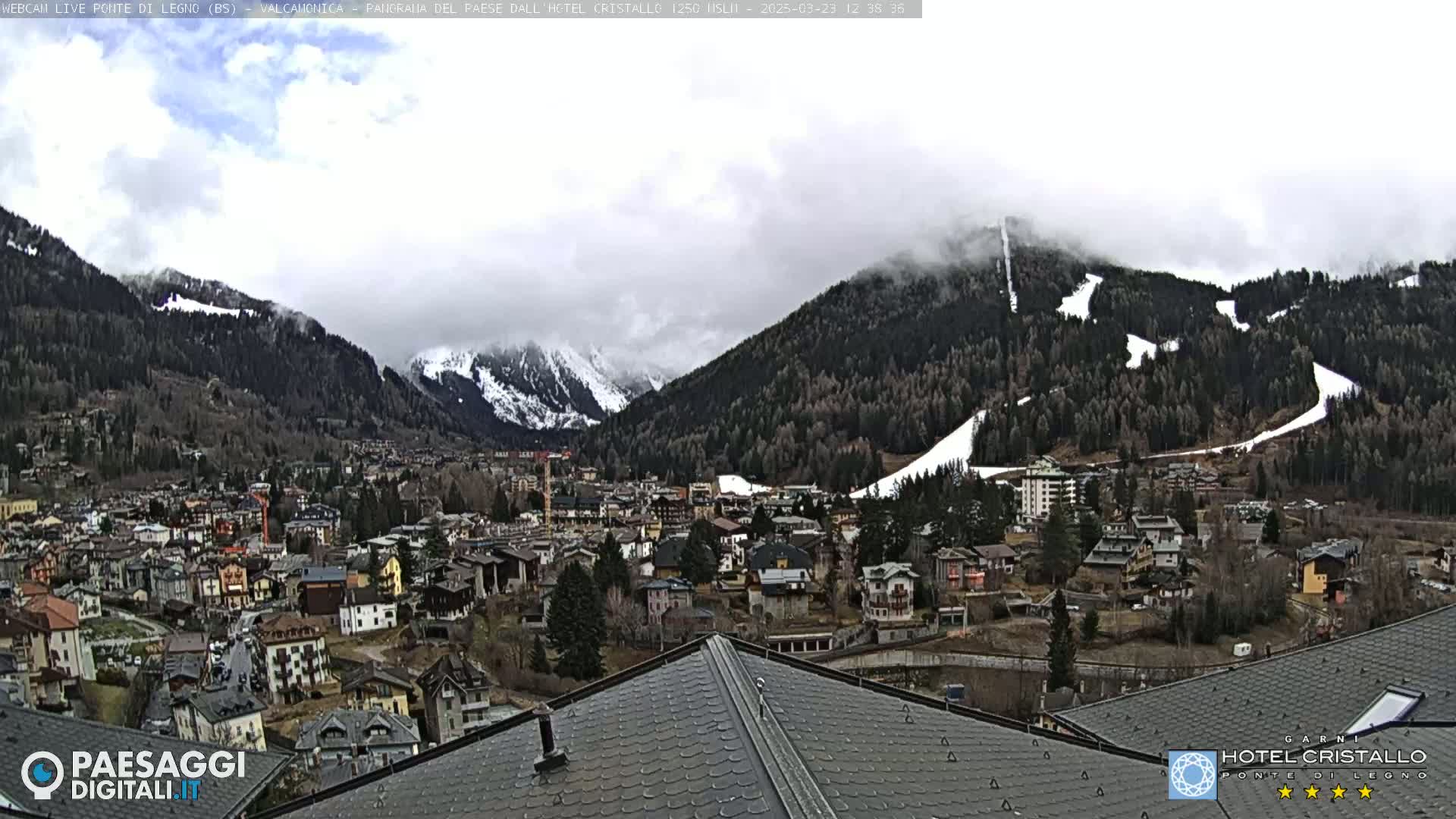 A mountain town nestled in a valley, partially obscured by clouds and fog, with snow-capped mountains in the background and ski runs visible on the slopes.