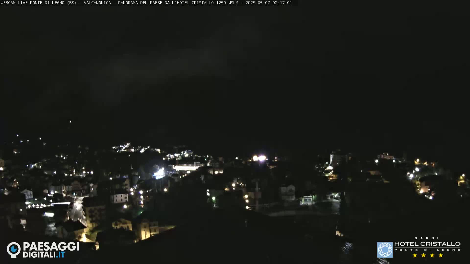 A nighttime, high-angle view of a town illuminated by scattered lights under a dark, cloudy sky.