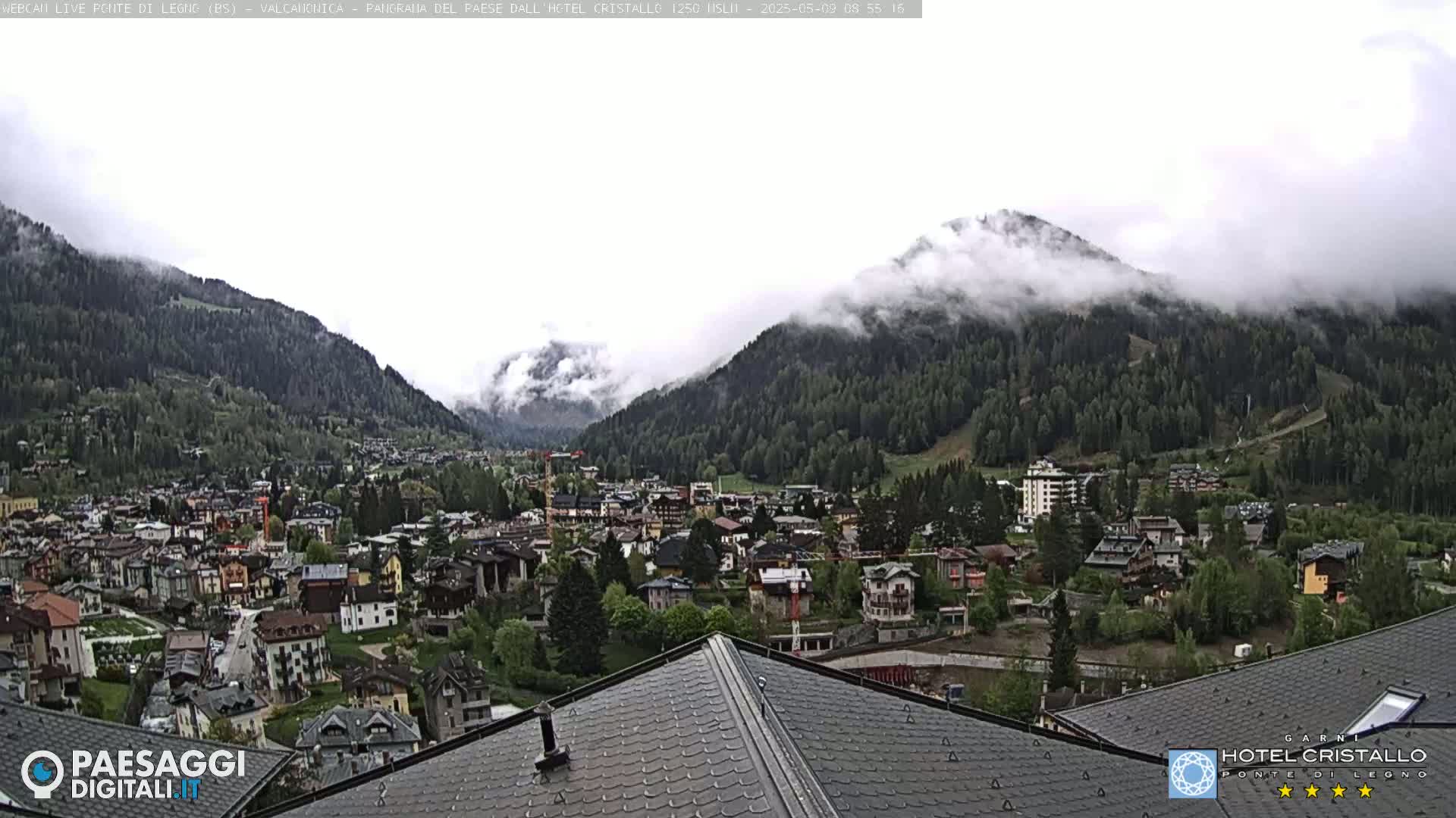A mountain town nestled in a valley, partially obscured by fog and low-hanging clouds, is viewed from a rooftop.