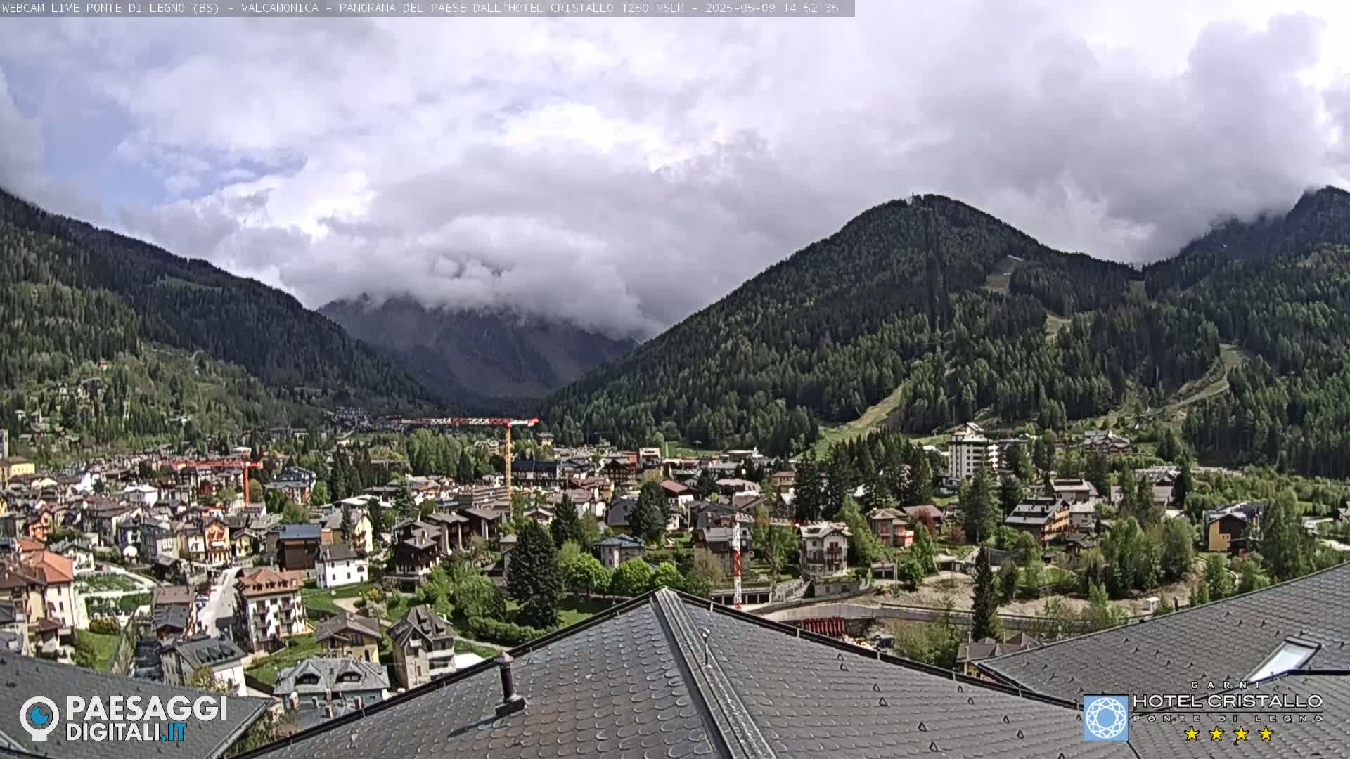 A partly cloudy day reveals a mountain valley town nestled amongst green hills and mountains, viewed from a rooftop.