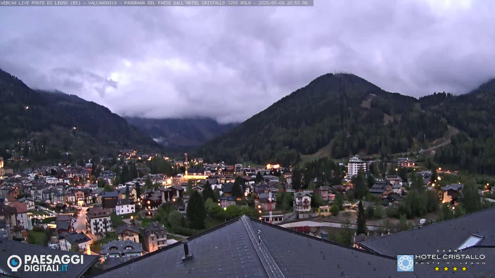 A nighttime view of a town nestled in a valley between mountains under a cloudy sky.