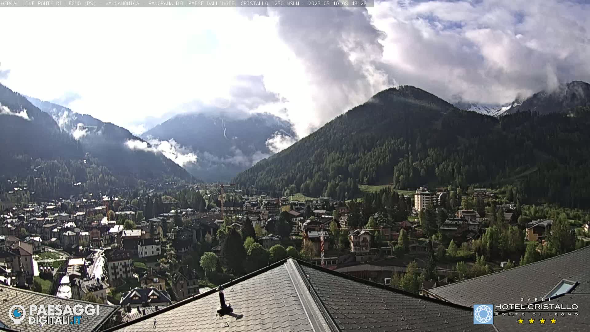 A mountain town nestled in a valley between forested mountains, partially obscured by clouds, is viewed from a rooftop.