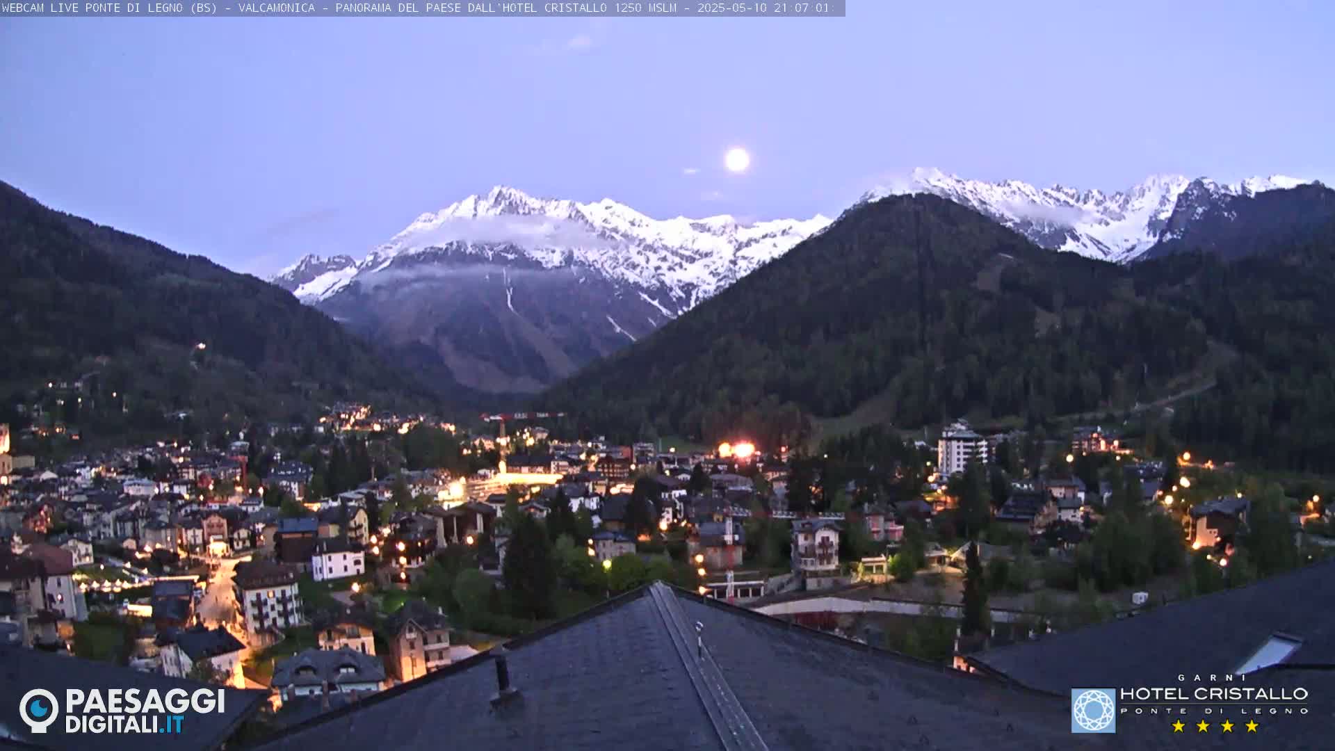 A nighttime view of a town nestled in a valley, illuminated by lights, with snow-capped mountains in the background under a twilight sky showing a full moon.