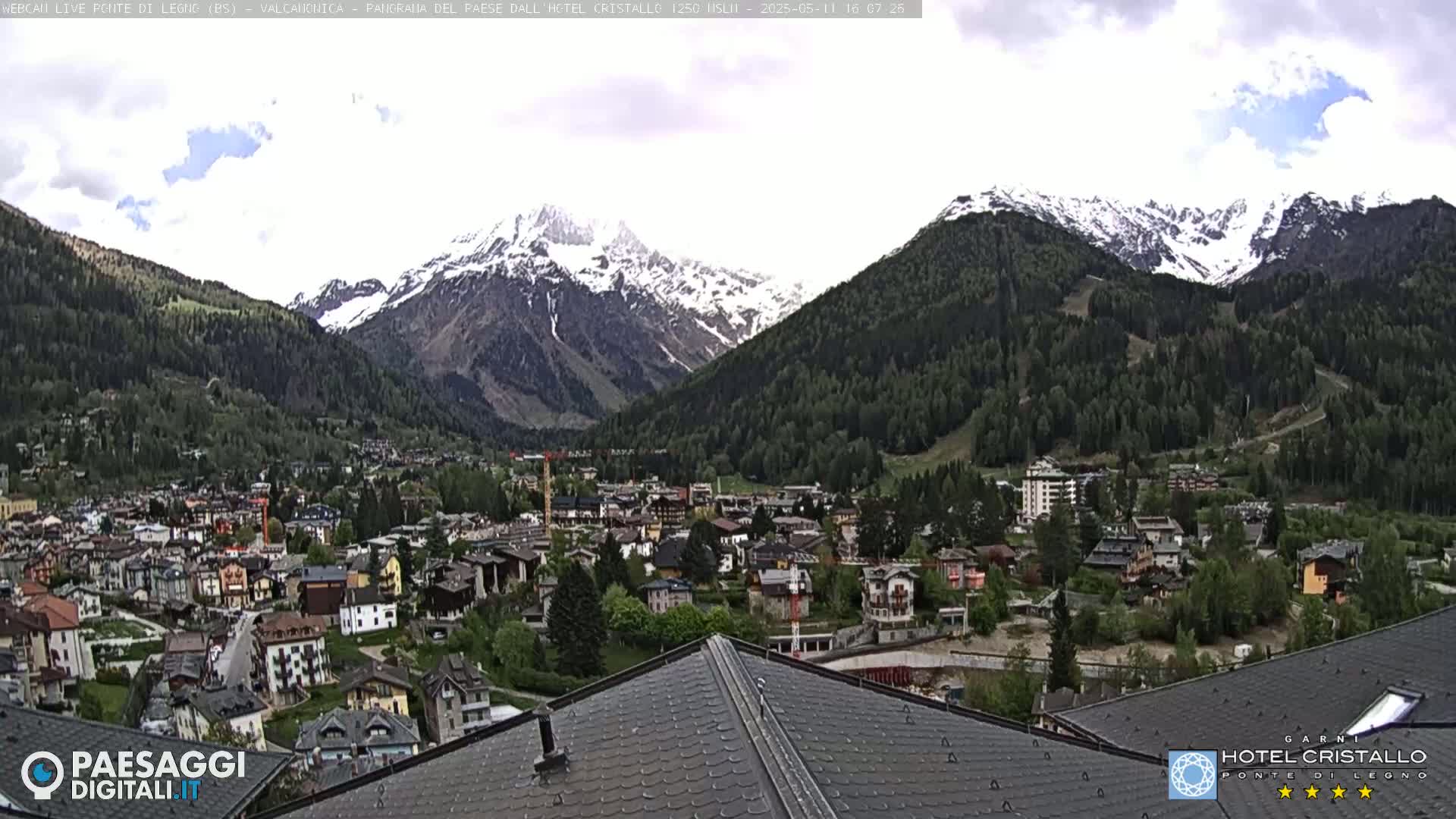 A mountain village nestled in a valley, with snow-capped mountains in the background under a partly cloudy sky.