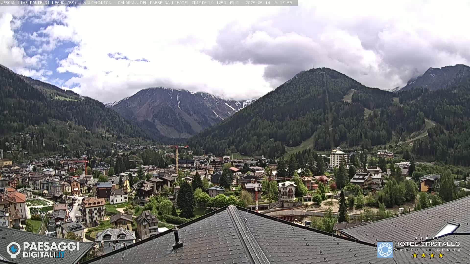 A partly cloudy day reveals a mountain valley town nestled amongst green hills and snow-capped mountains.