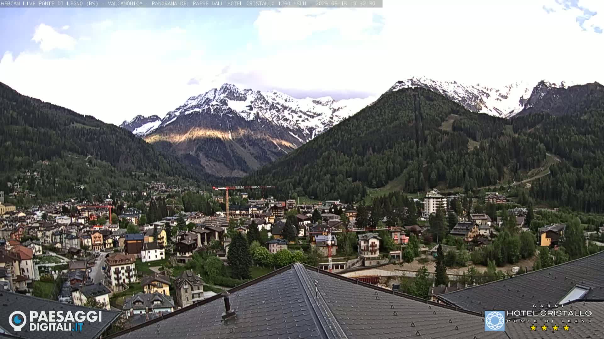 A mountain village nestled in a valley, with snow-capped mountains in the background under partly cloudy skies.