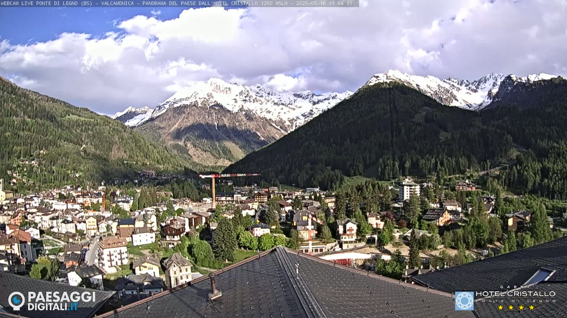 A partly cloudy day reveals a mountain town nestled in a valley surrounded by snow-capped mountains.