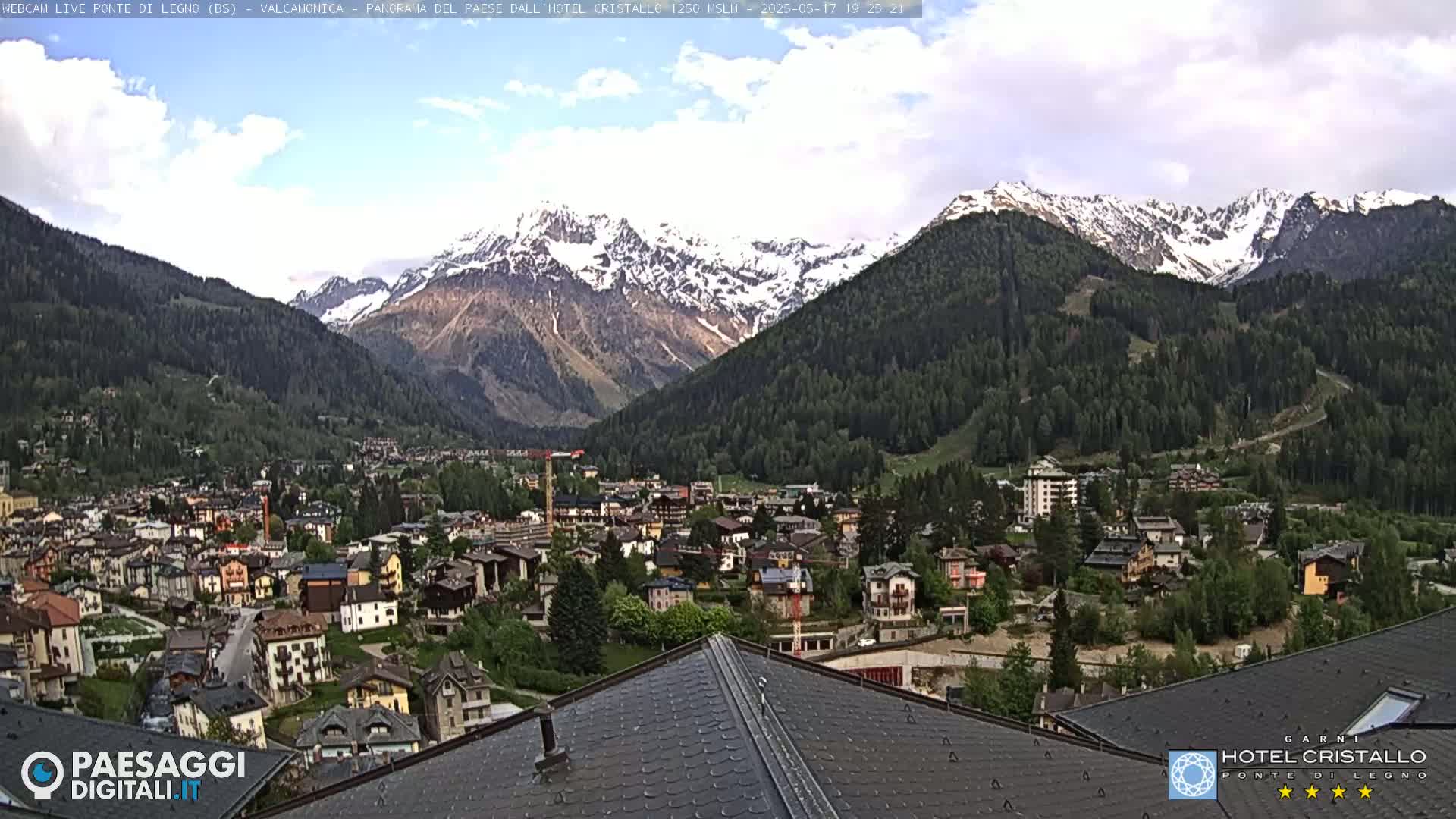 A partly cloudy day reveals a mountain village nestled in a valley, with snow-capped mountains in the background.