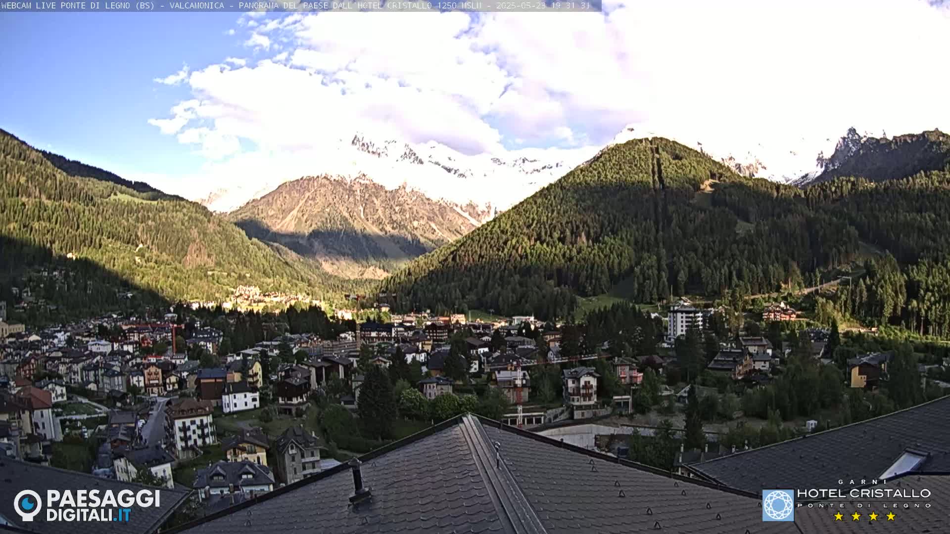 A partly cloudy day reveals a mountain village nestled in a valley, with snow-capped mountains rising in the background.