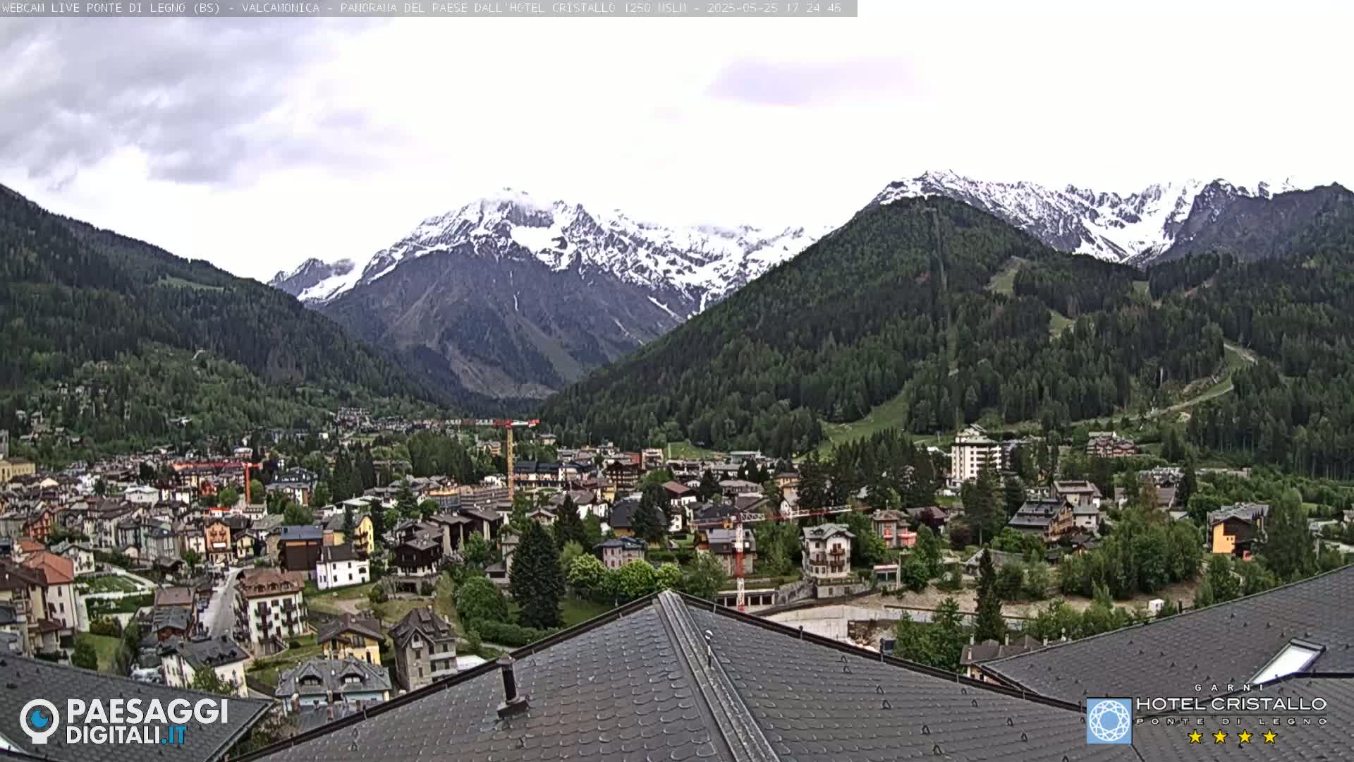 A mountain town nestled in a valley, with snow-capped mountains in the background under a partly cloudy sky.