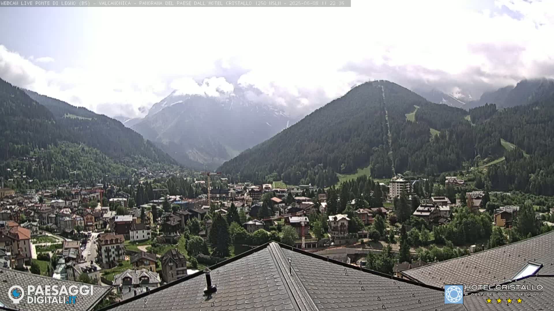 A partly cloudy mountain valley town is viewed from a rooftop, with green hills and distant snow-capped mountains in the background.