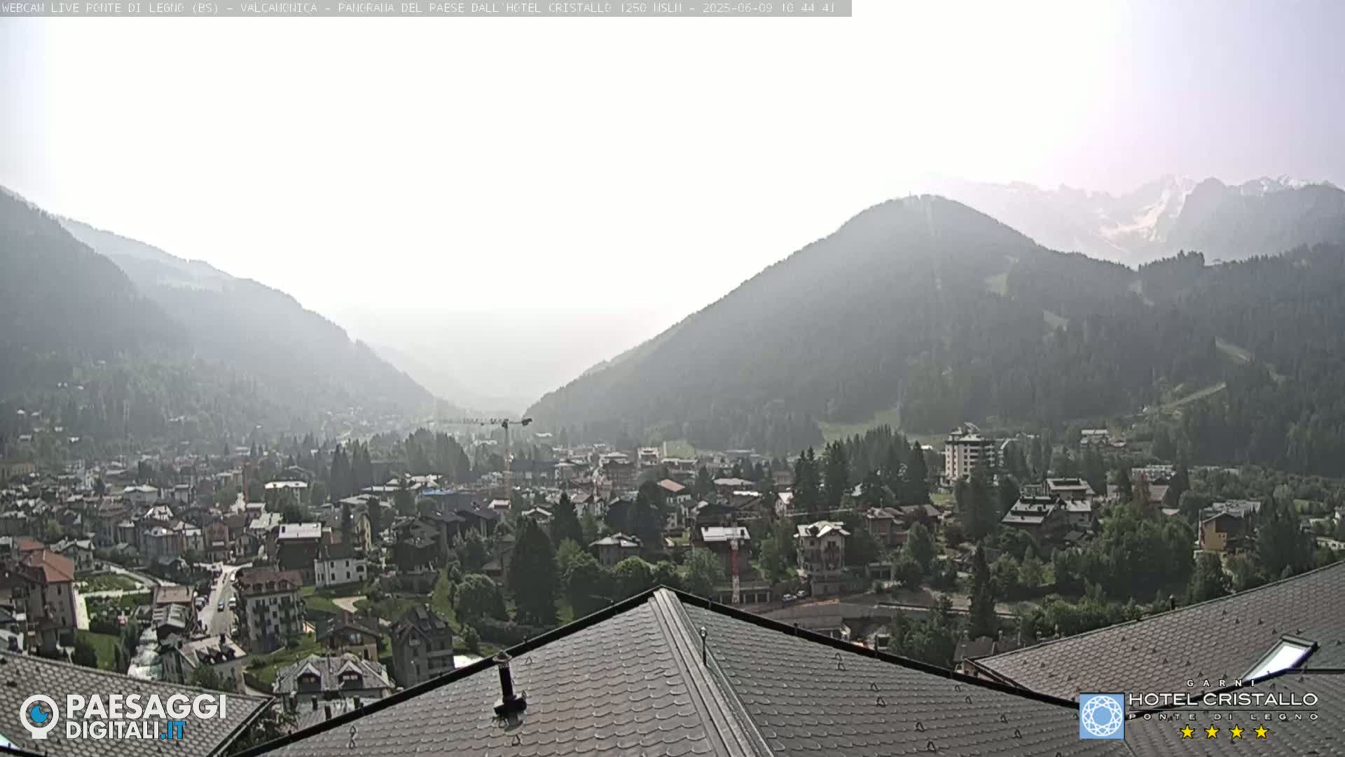 A hazy view from a mountaintop shows a valley town nestled among hills, with distant mountains partially obscured by atmospheric conditions.