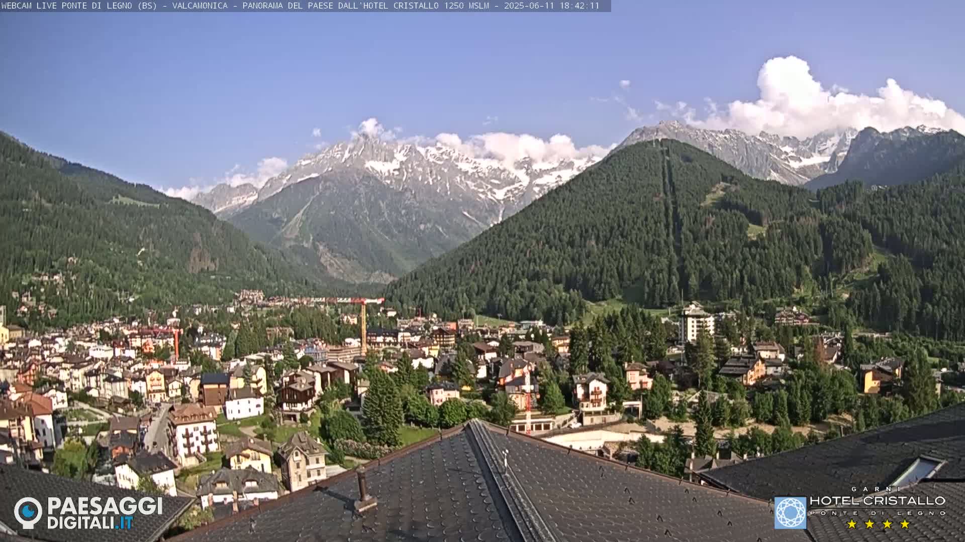 A partly cloudy day reveals a mountain town nestled in a valley, with snow-capped mountains in the background.