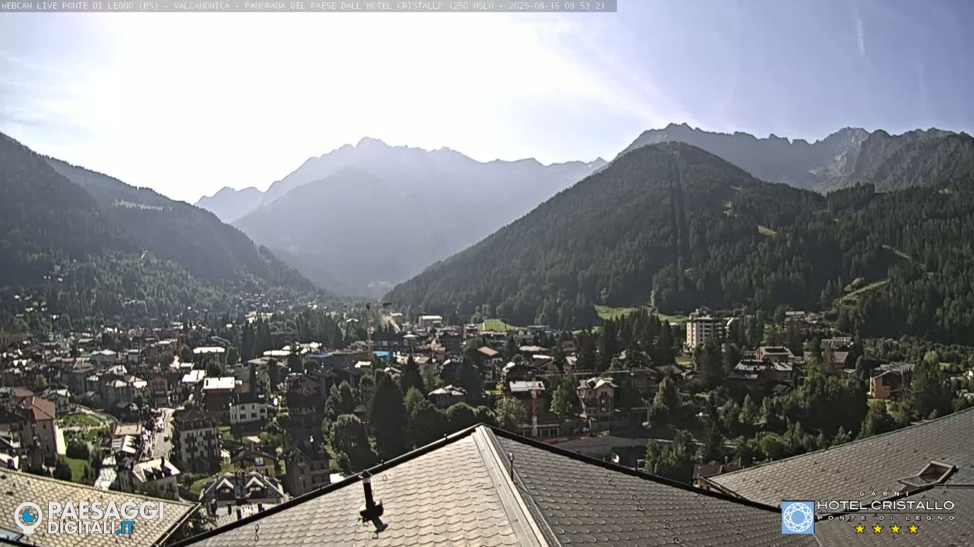 A mountain valley town is viewed from a rooftop on a sunny day, with a range of mountains in the background.