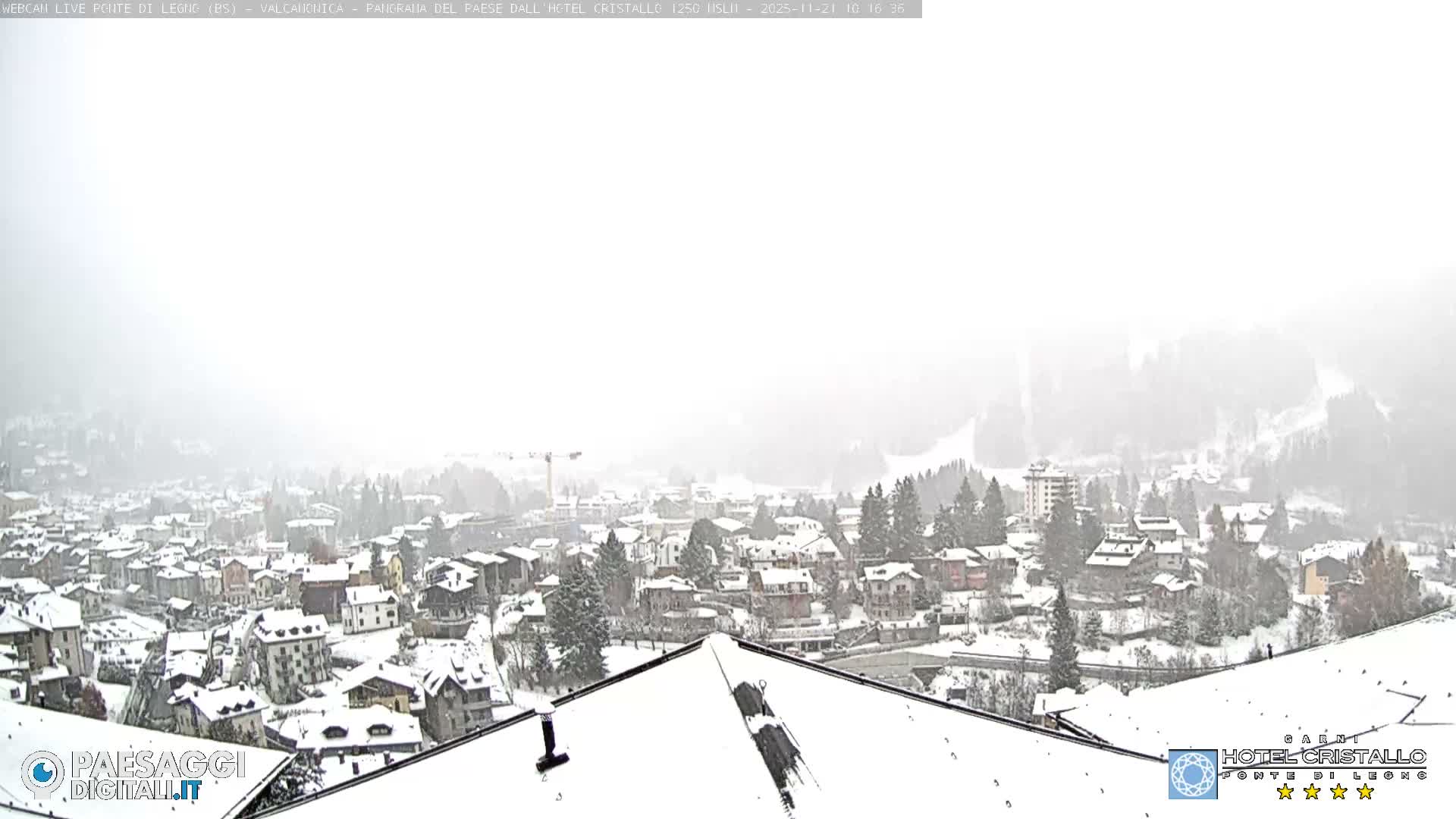A panoramic view shows a snow-covered village nestled among frosted coniferous trees and partially obscured mountains under a bright, overcast, and snowy sky.