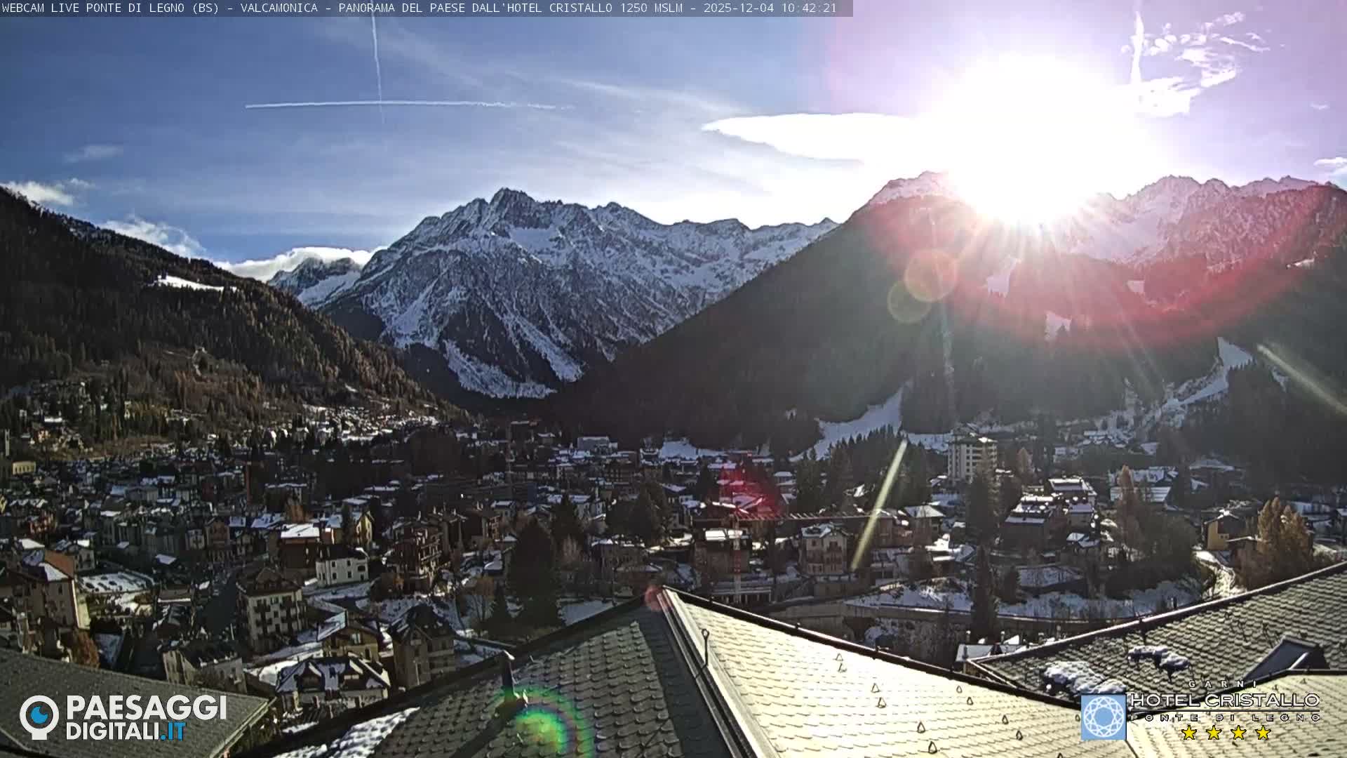A bright sun flares over a snow-dusted mountain village nestled in a valley, surrounded by snow-capped peaks under a clear, sunny sky.