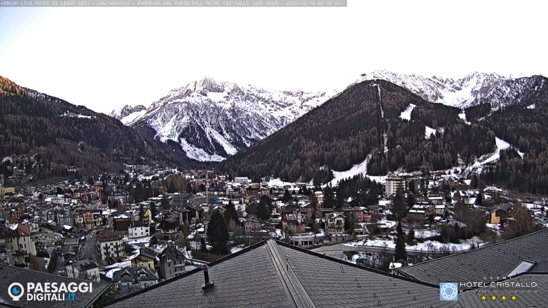 A bright sun flares over a snow-dusted mountain village nestled in a valley, surrounded by snow-capped peaks under a clear, sunny sky.
