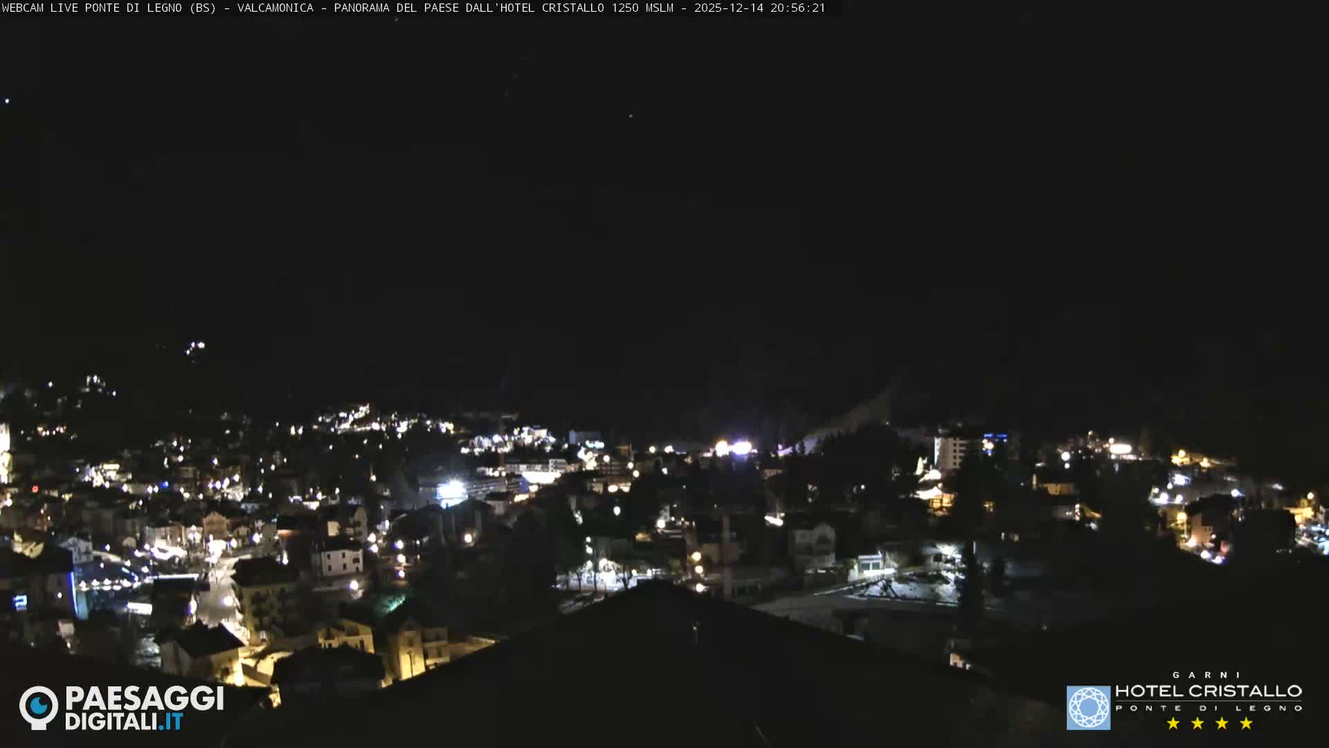 A bright sun flares over a snow-dusted mountain village nestled in a valley, surrounded by snow-capped peaks under a clear, sunny sky.