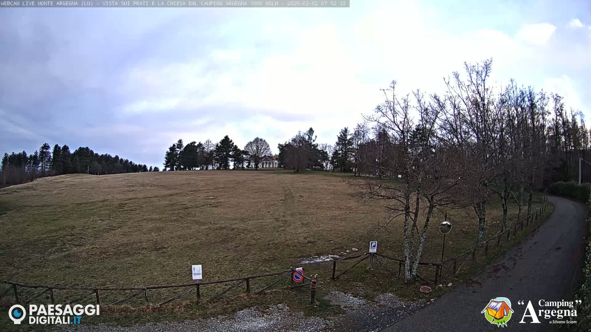 A mostly cloudy day reveals a large, open grassy field bordered by leafless trees and a paved road leading to a wooden fence.