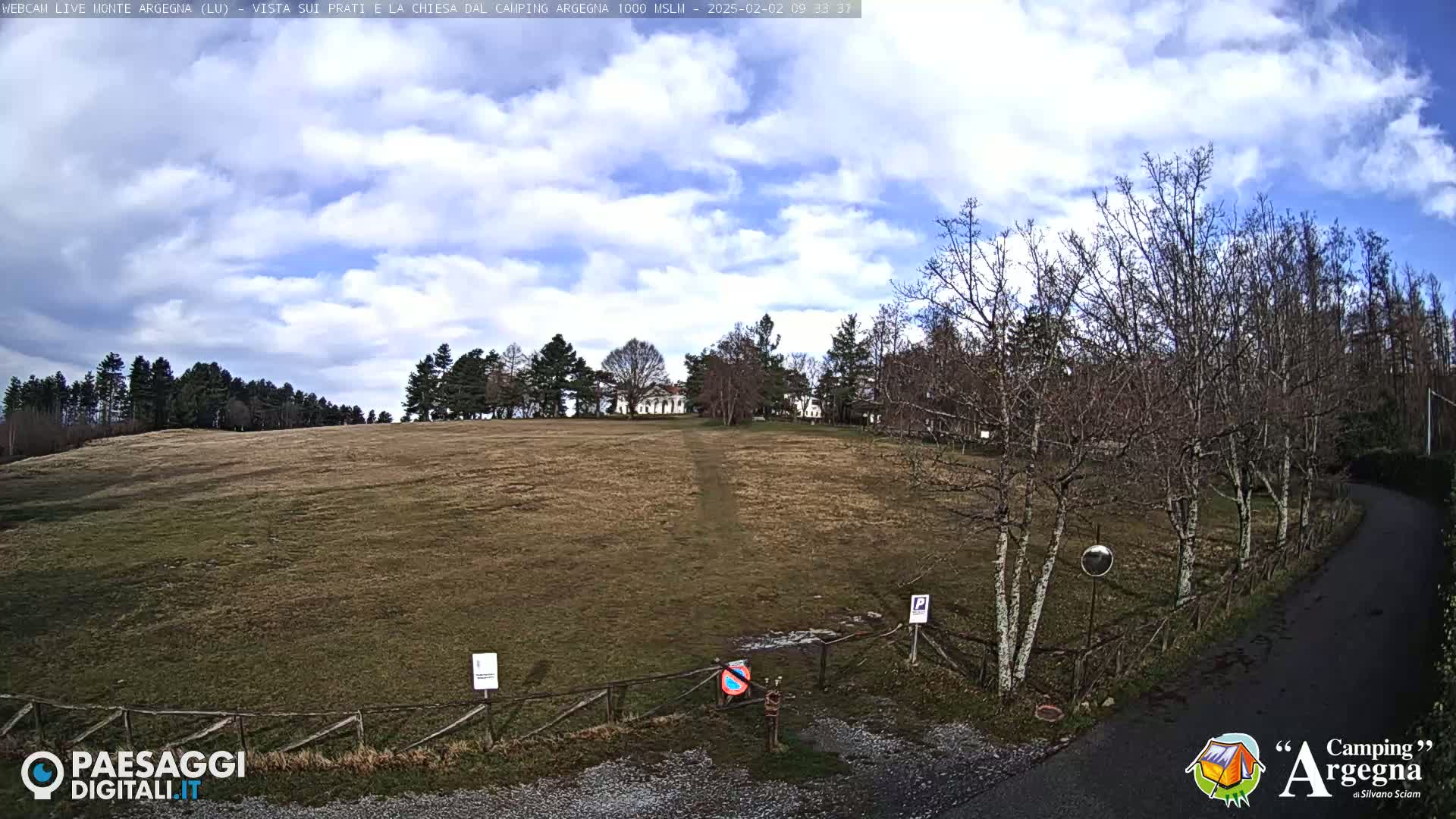 A partly cloudy day reveals a brown grassy field, a dirt road, leafless trees, and a building in the distance.