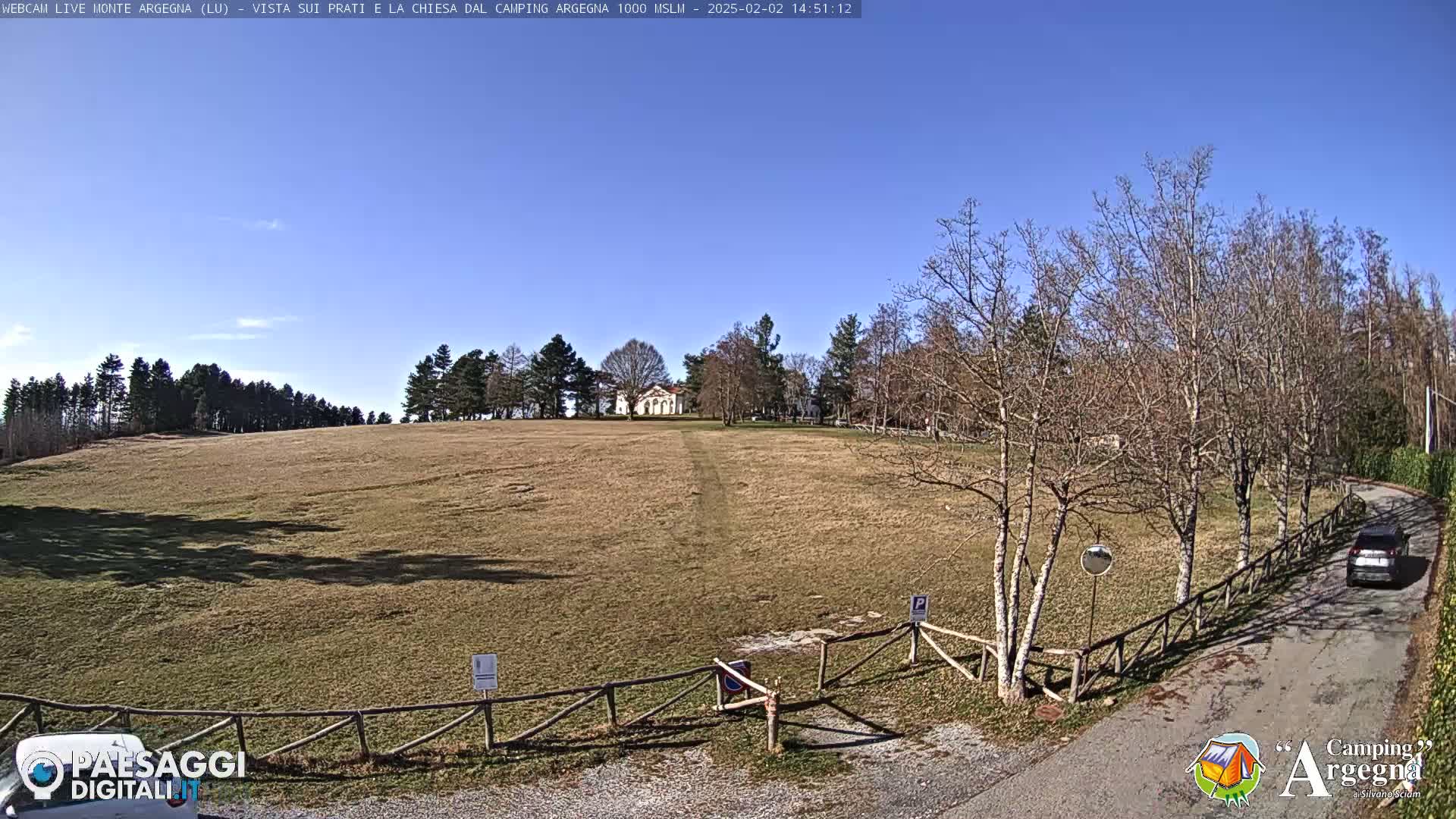 A large, grassy field slopes gently upward toward a small white building, under a clear blue sky with leafless trees lining a road to the right.