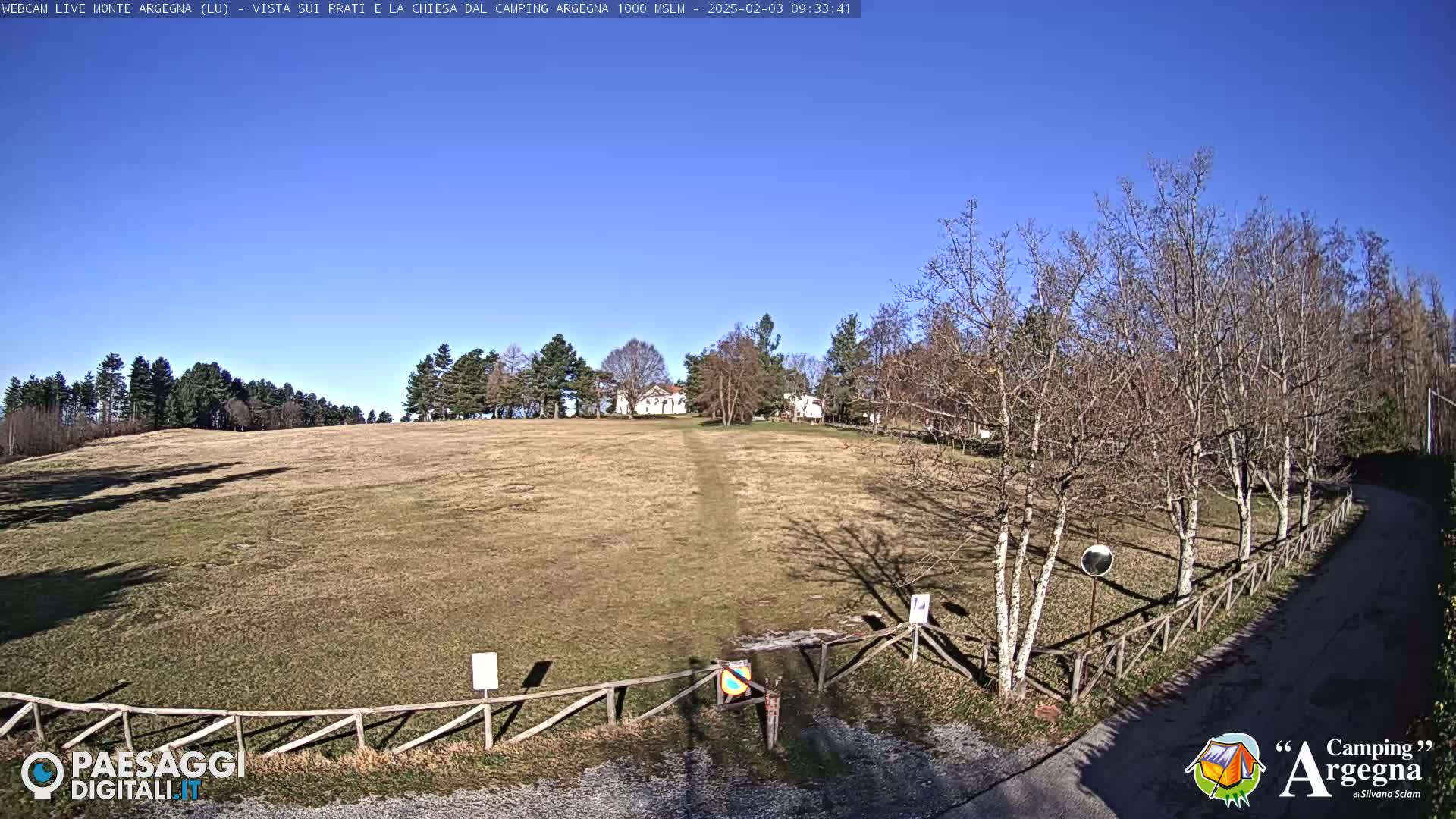 A grassy field stretches toward a small building and trees under a clear blue sky.