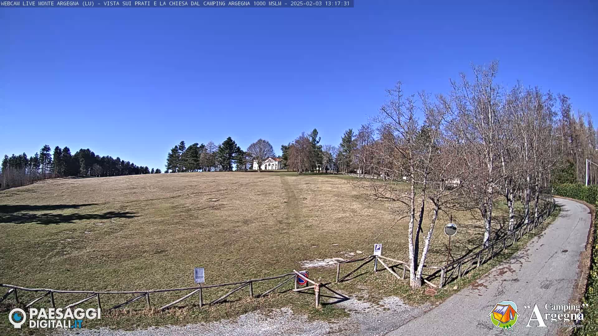 A large, grassy field slopes gently upward toward a small white building, under a clear blue sky, with leafless trees lining a paved road to the right.