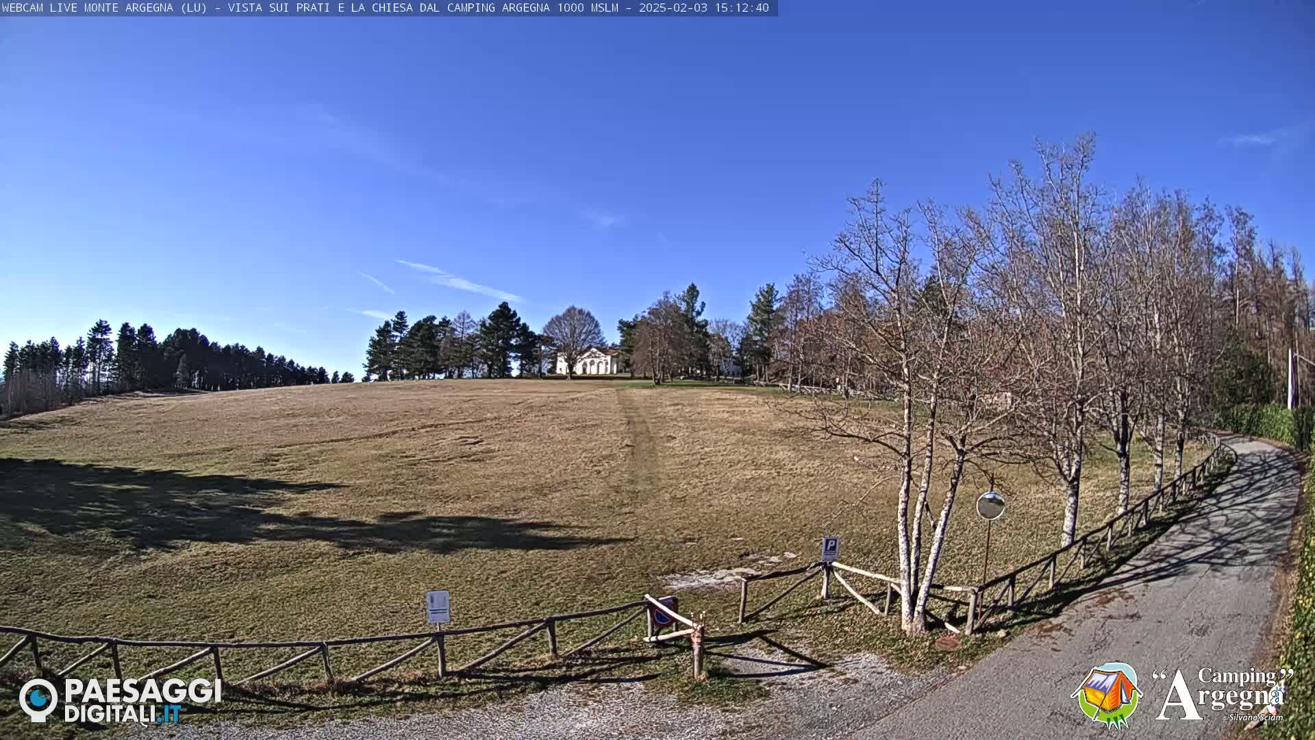 A wide, brown grassy field stretches towards a small white building under a clear blue sky, with leafless trees lining a road to the right.