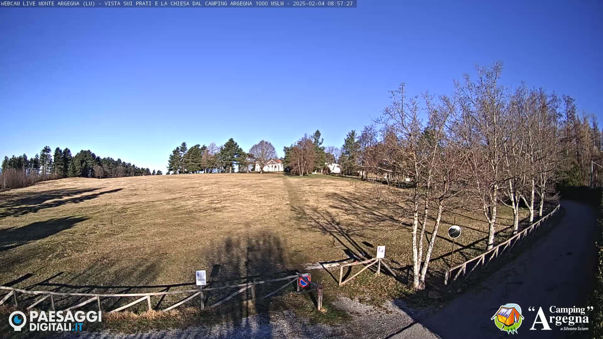 A tan grassy field stretches toward a small white building, bordered by leafless trees under a clear blue sky.