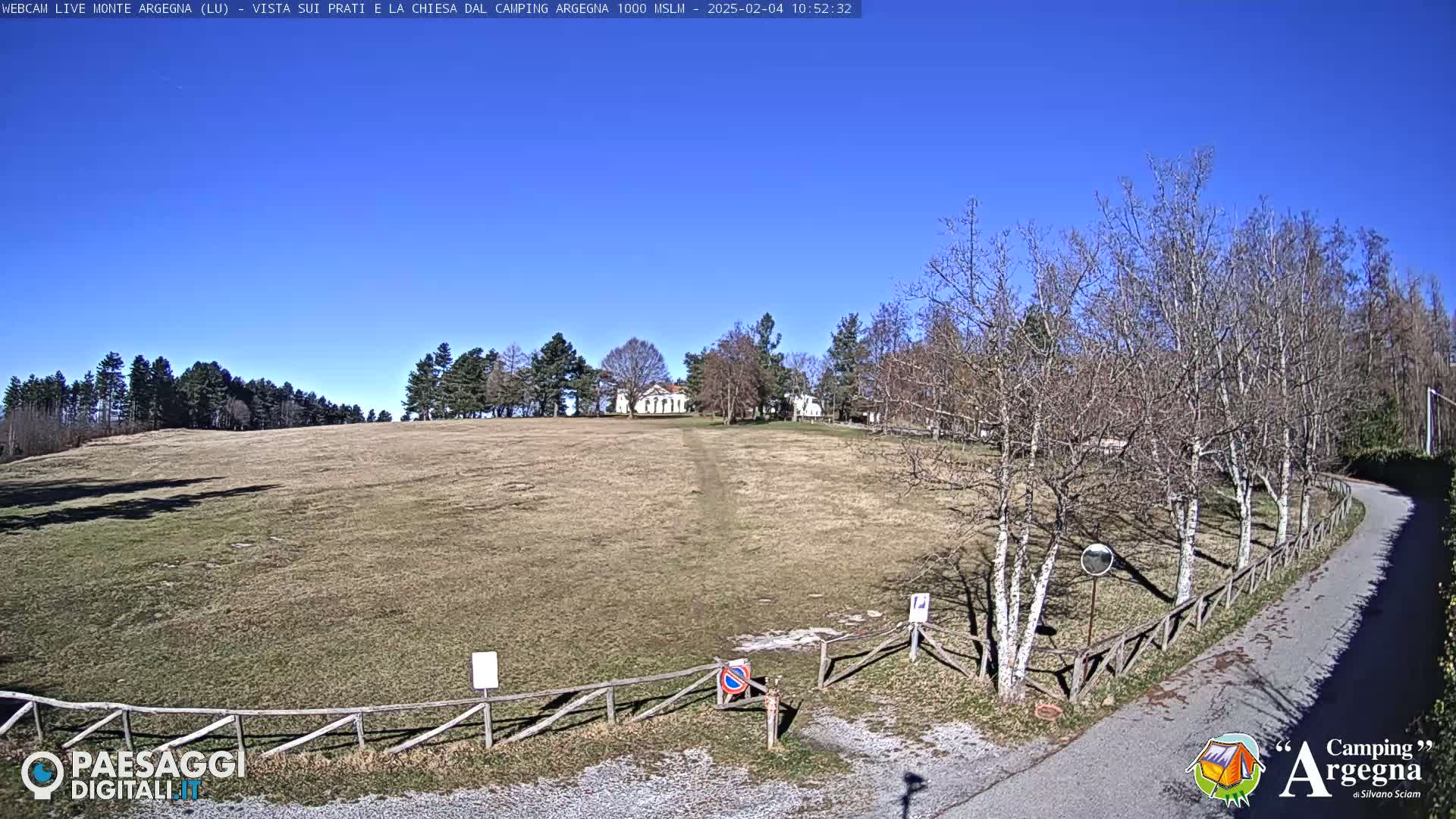 A large, grassy field with a white building in the distance under a clear blue sky is seen from a slight uphill angle, with bare trees lining a paved road to the right.
