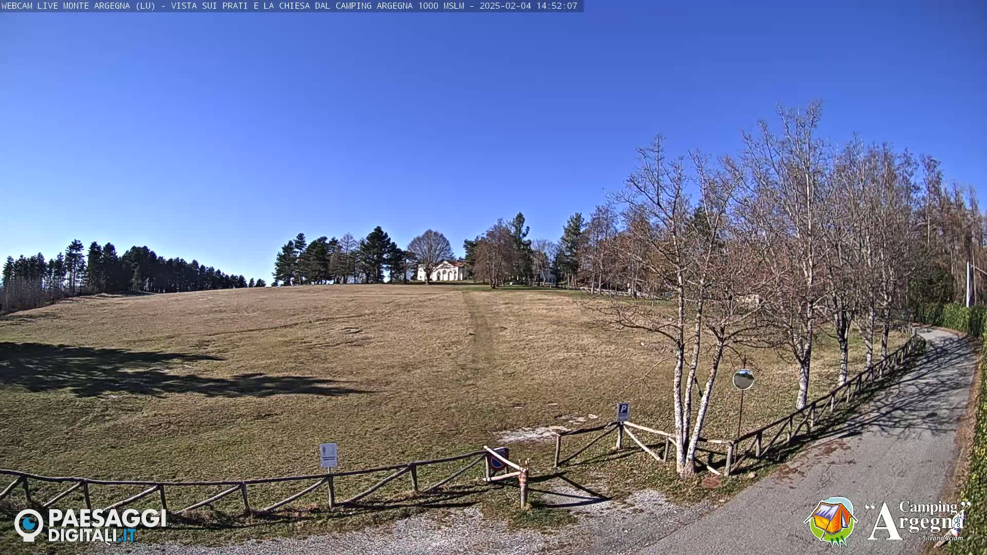 A wide, grassy field stretches towards a small, white building under a clear blue sky, with leafless trees lining a road to the right.