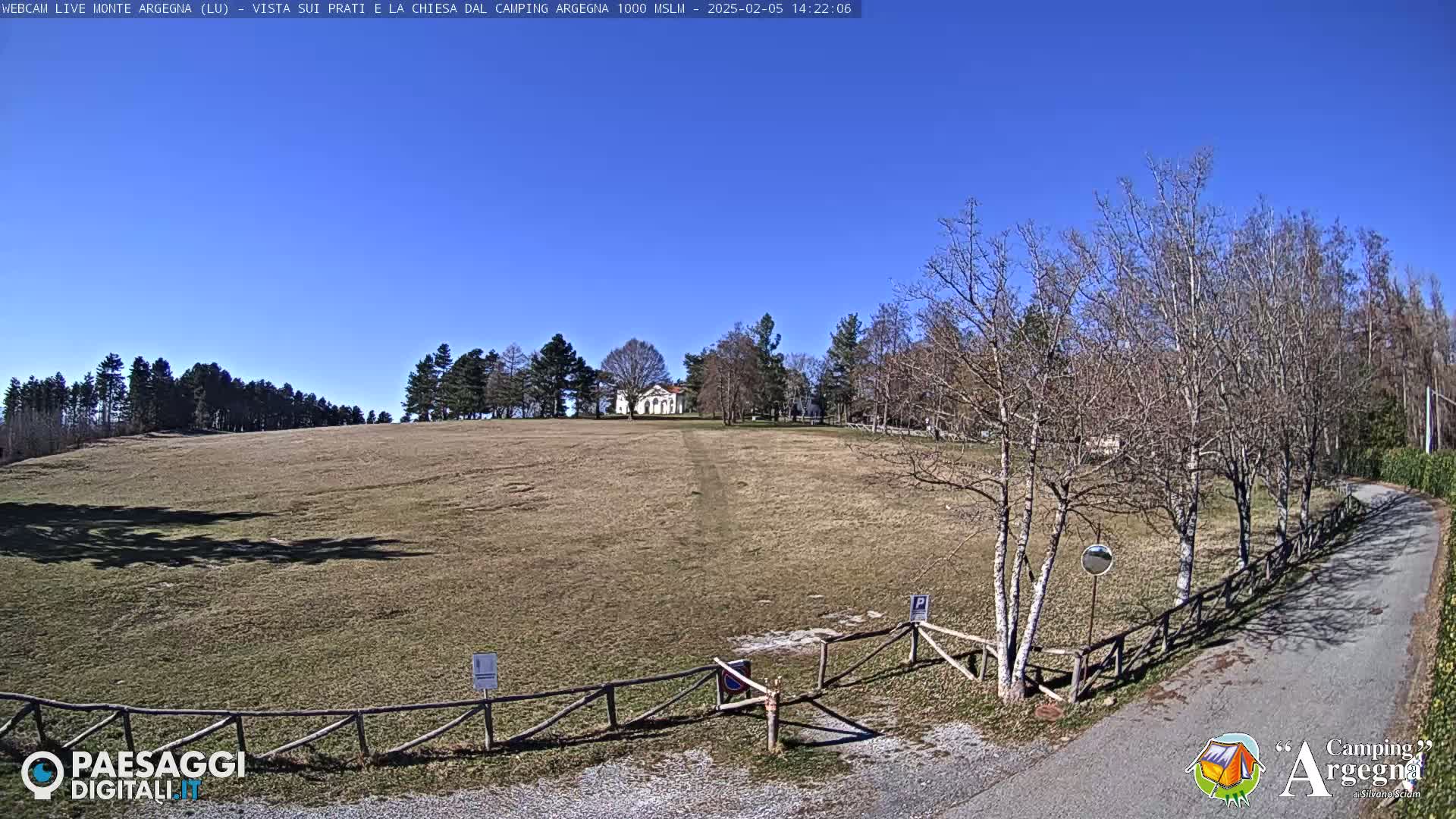 A wide, brown field stretches toward a small white building under a clear blue sky, with bare trees lining a nearby road.