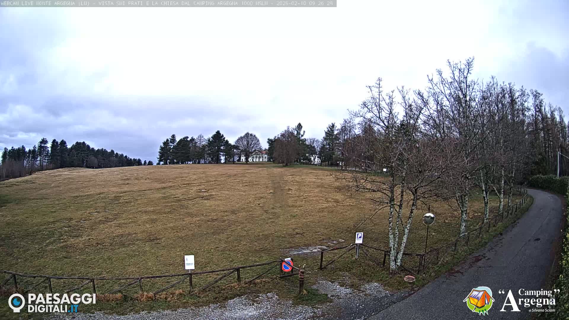 A mostly cloudy day reveals a view of a brown field, a distant building, and bare winter trees lining a paved road.