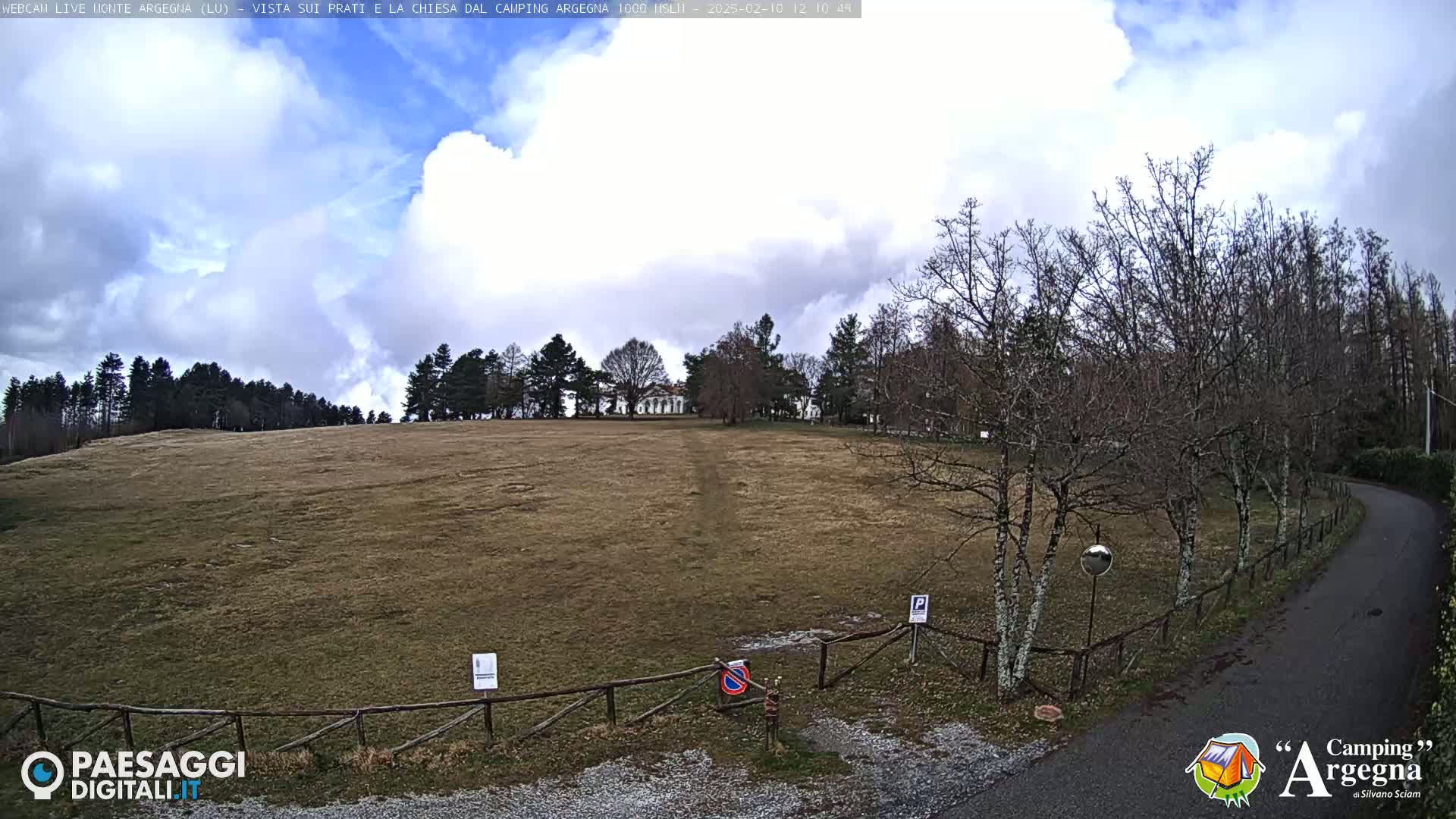 A wide, grassy field slopes gently downward toward a paved road bordered by leafless trees under a partly cloudy sky.