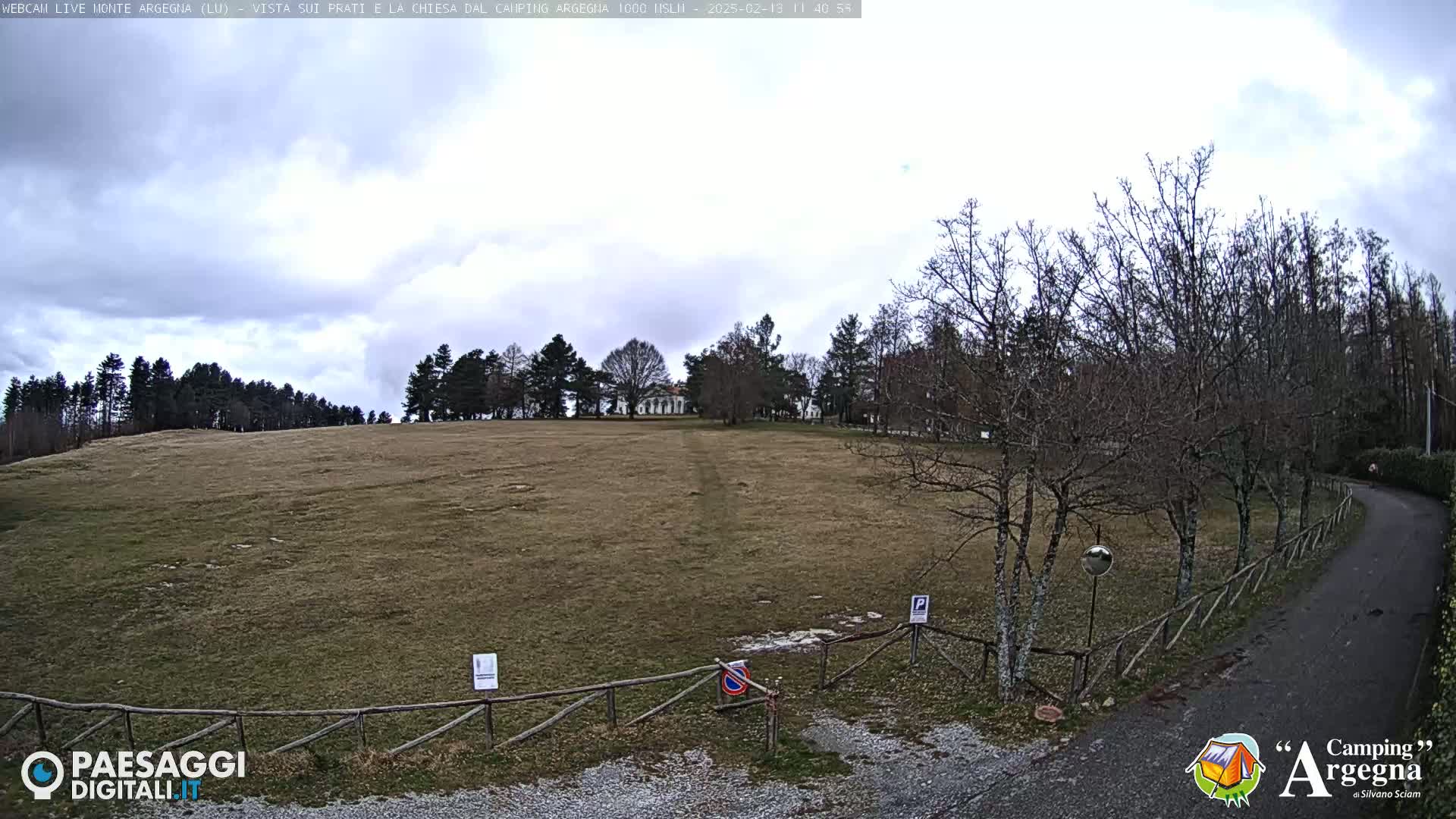 A wide, flat, grassy field with leafless trees and a small building in the distance under a mostly cloudy sky.
