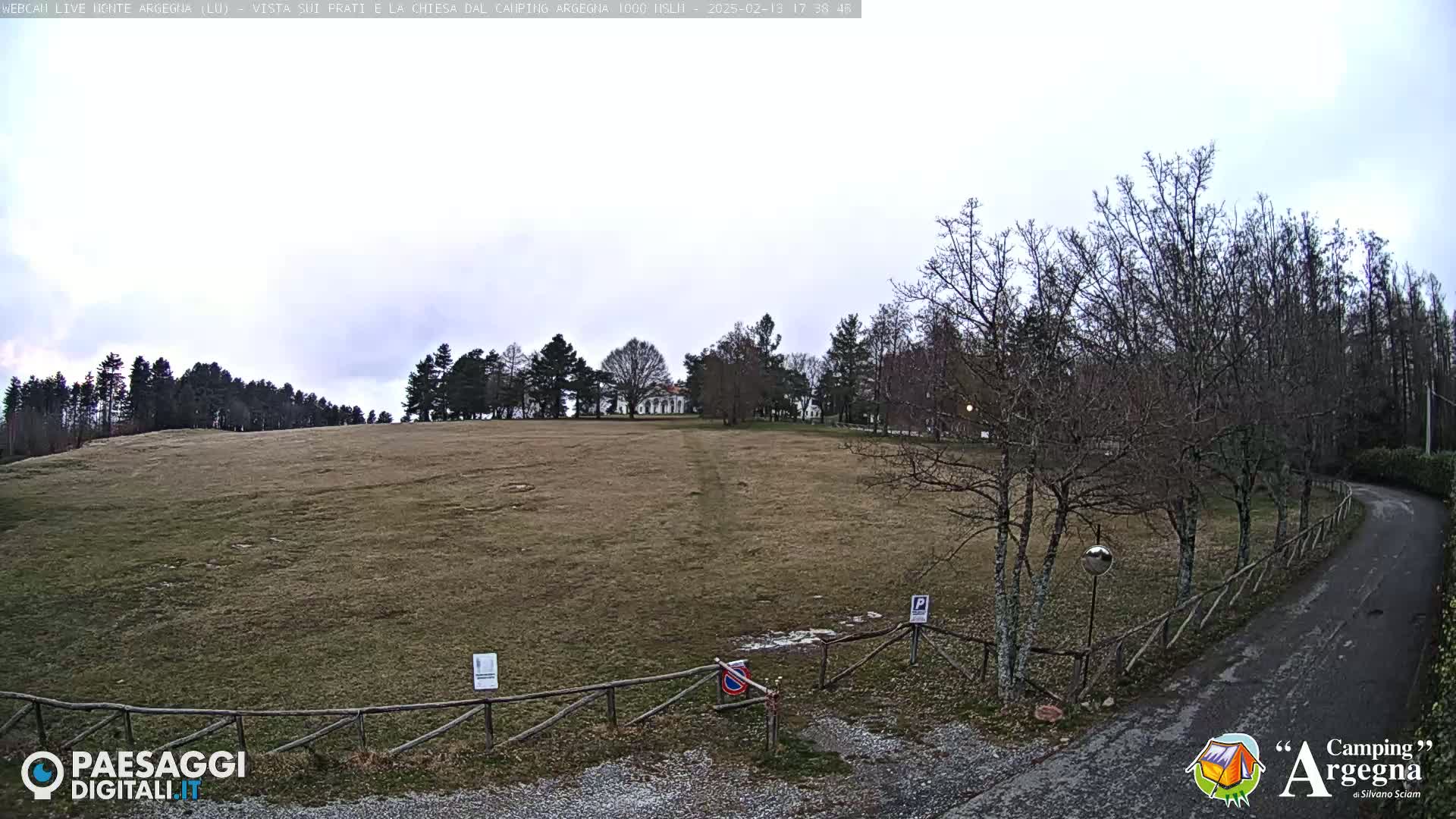 A grassy field, bordered by leafless trees and a wooden fence, leads to a building in the distance under an overcast sky.