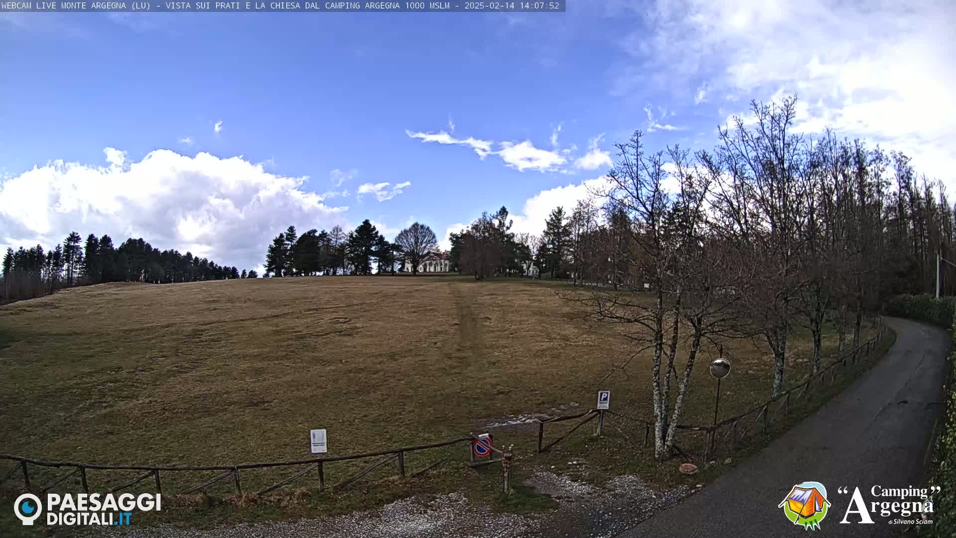 A mostly sunny day with partly cloudy skies shows a wide, grassy field bordered by leafless trees and a curving roadway.