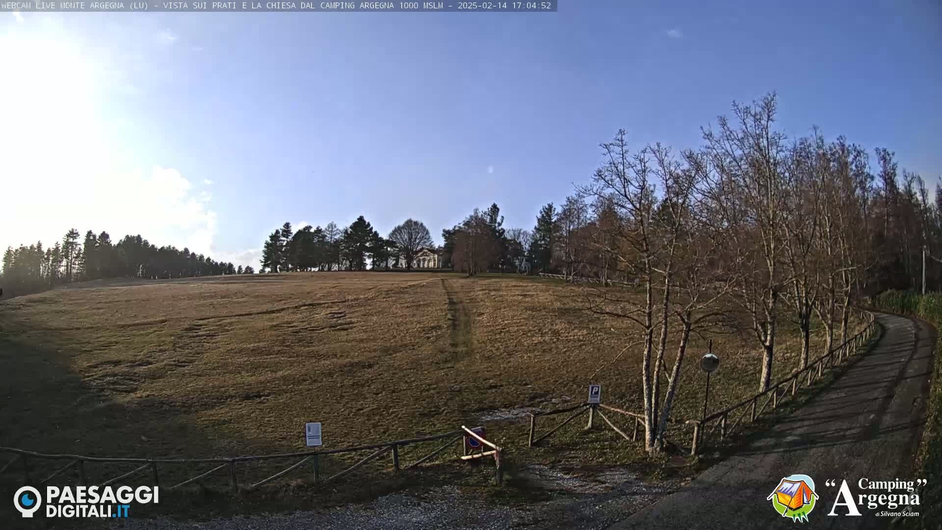 A wide, brown field stretches towards a small white building nestled among bare trees under a clear blue sky.