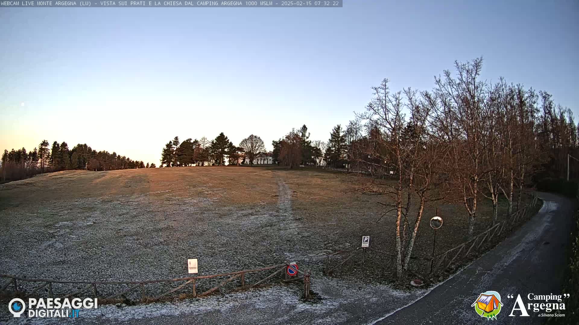 A frost-covered field is visible beyond a wooden fence, with leafless trees and a paved road leading to the right under a clear, pale blue sky.