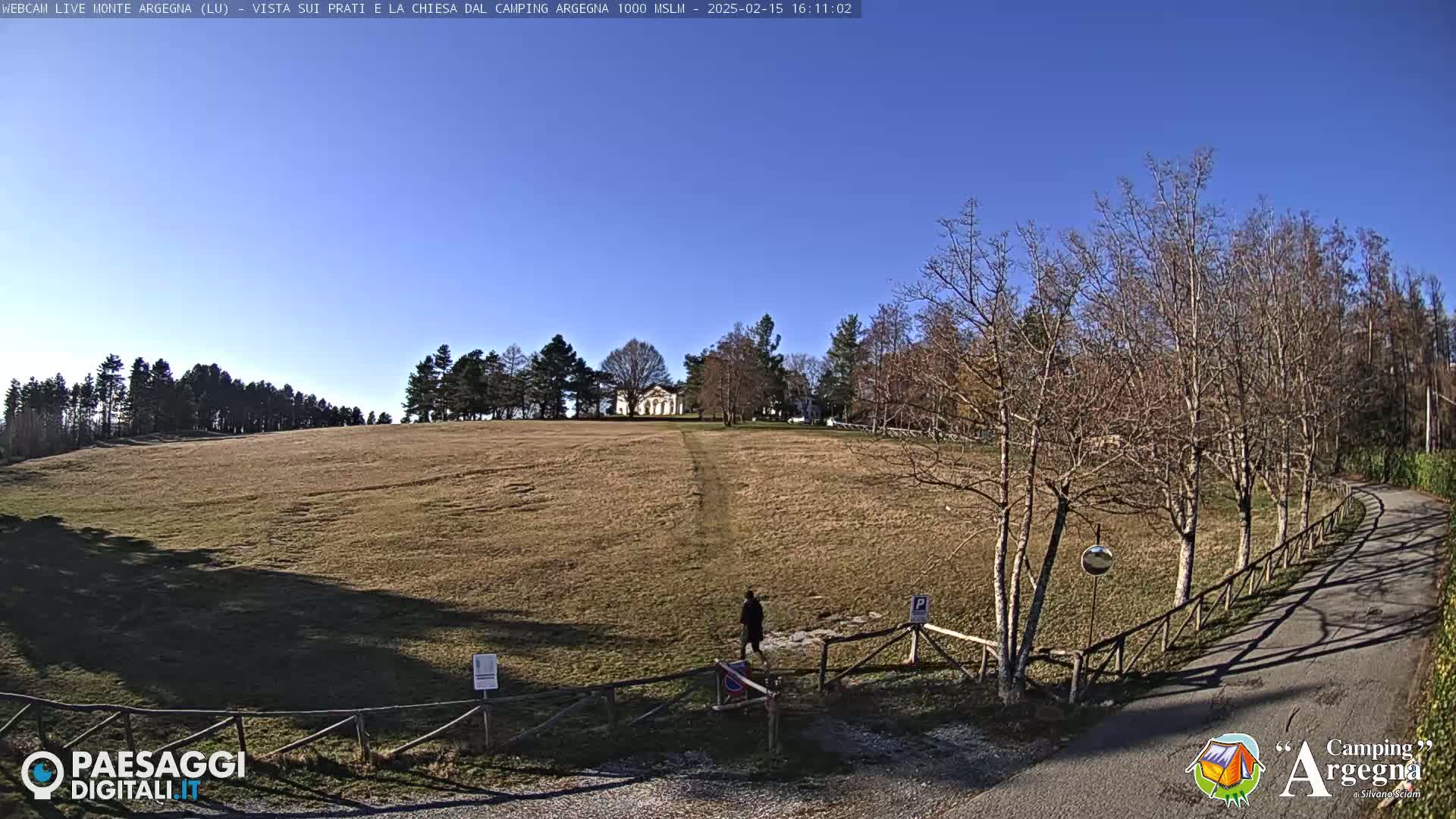 A person walks toward a white building on a hill in a field with leafless trees and a clear blue sky.