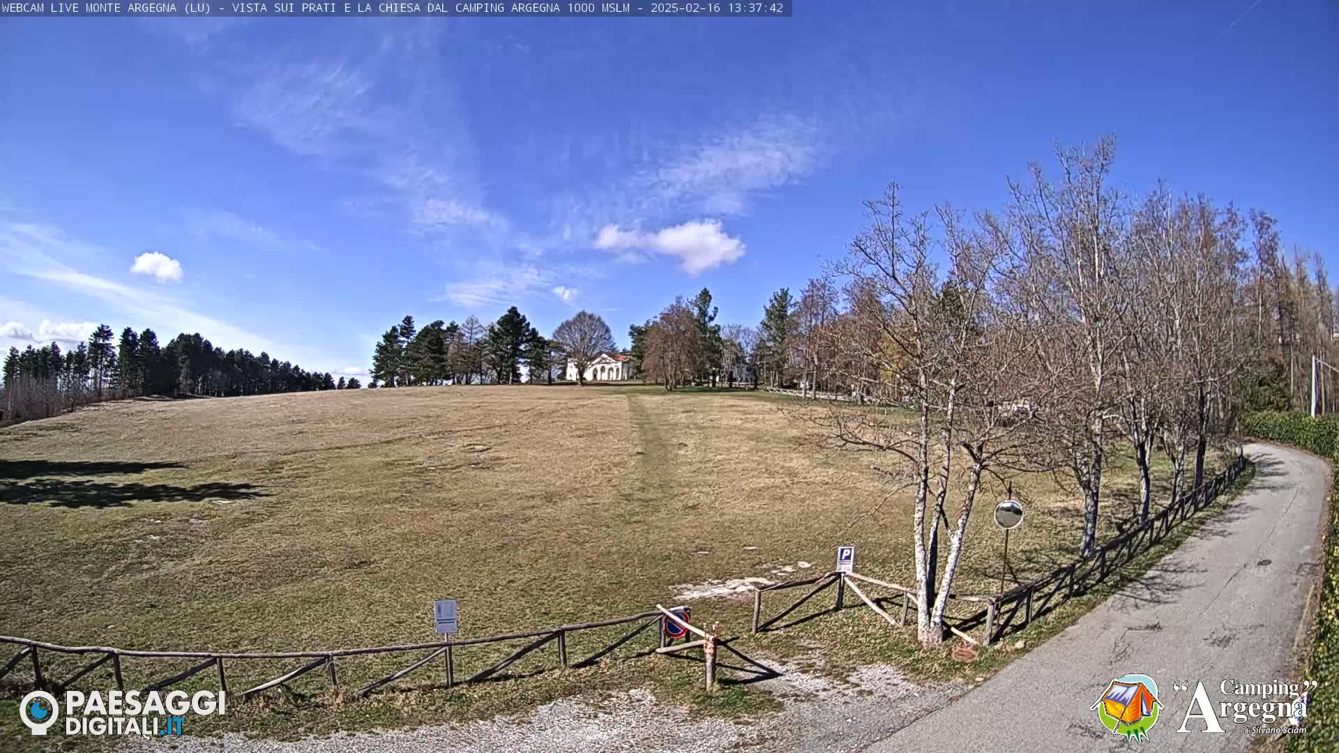A wide, grassy field slopes gently down to a road, with a small building visible in the distance under a mostly clear blue sky.