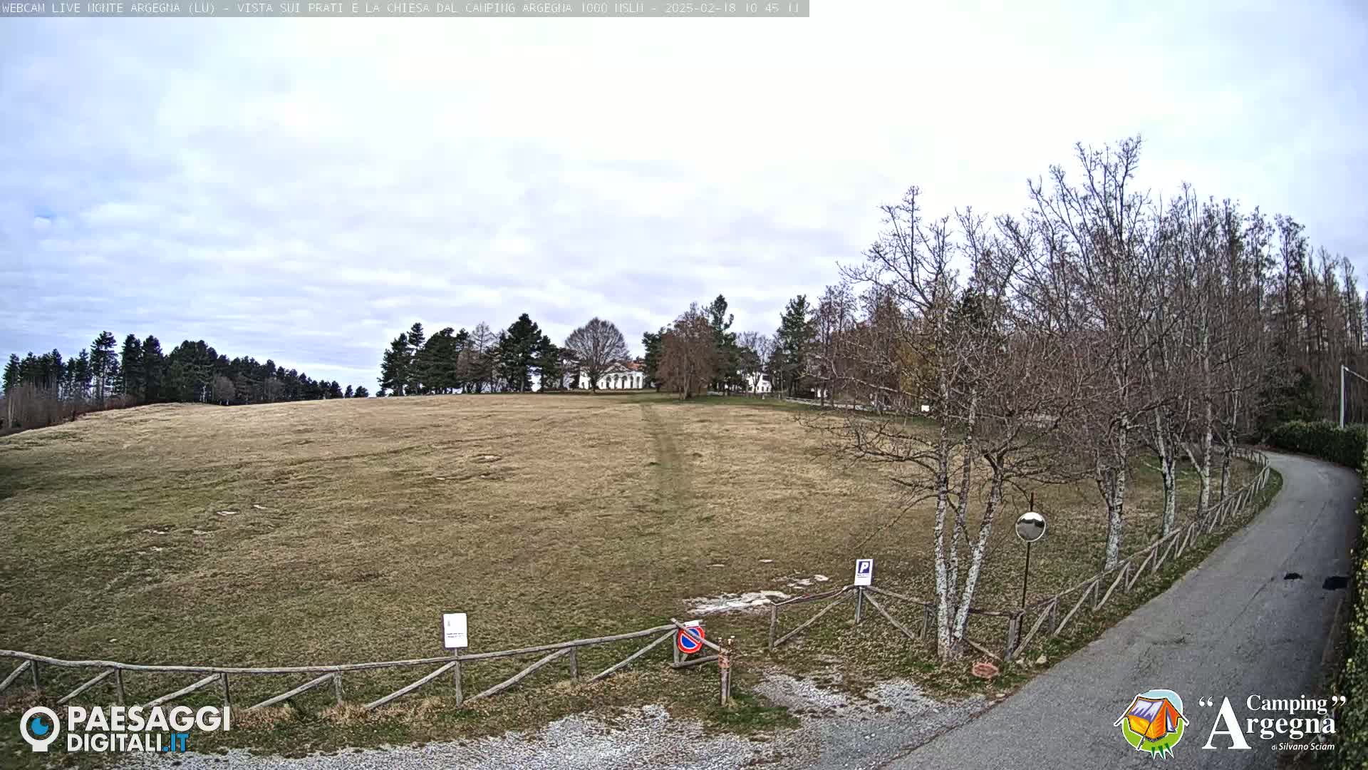 A wide, grassy field slopes gently upward toward a building nestled among bare winter trees under a mostly cloudy sky.
