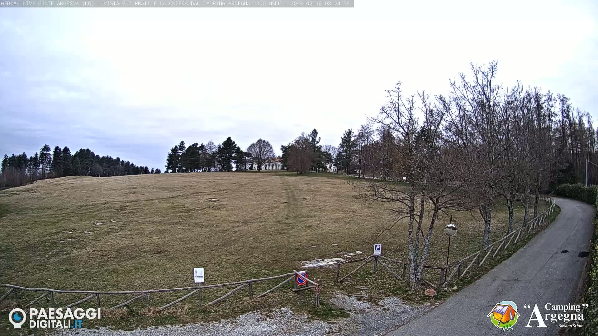 A wide, brown field stretches to a line of bare trees and a building in the distance under a cloudy sky.