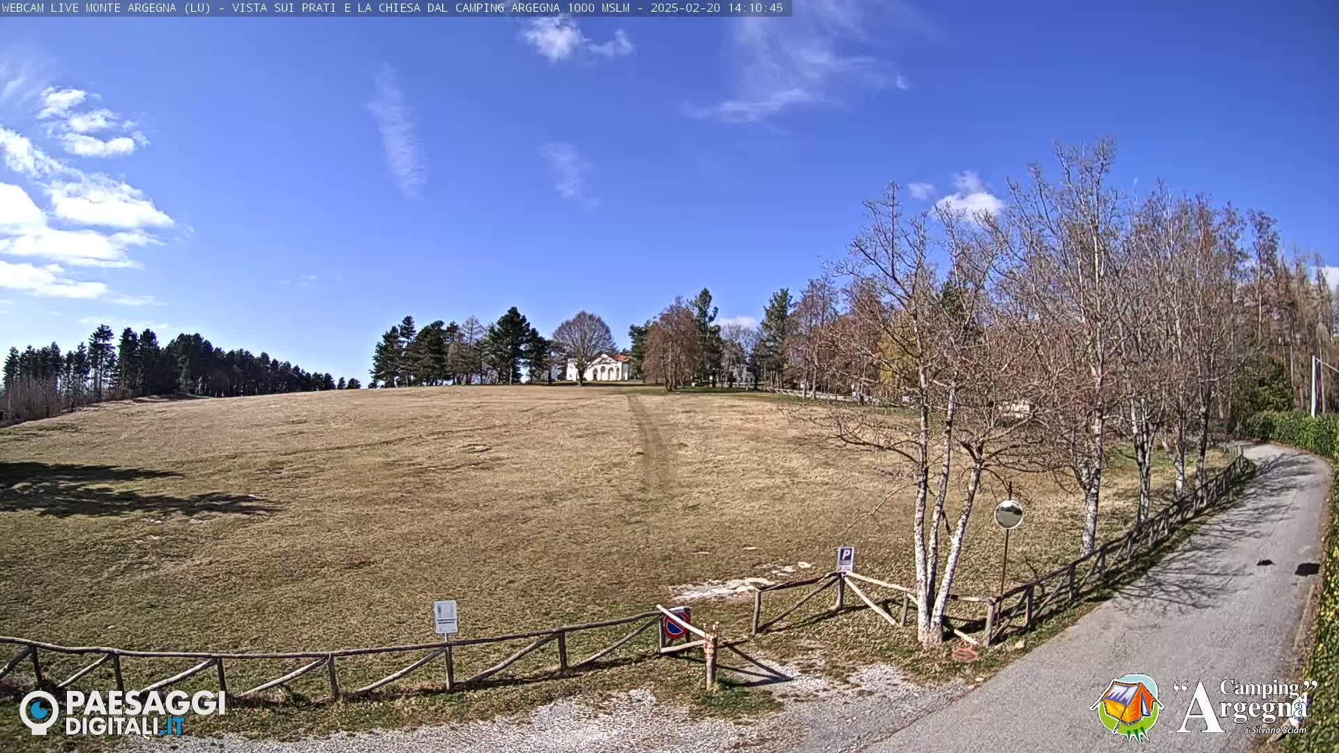 A large, flat, brown field stretches toward a small white building under a mostly sunny, blue sky with a few scattered clouds; bare trees line the edges of the field and a paved road is visible in the foreground.