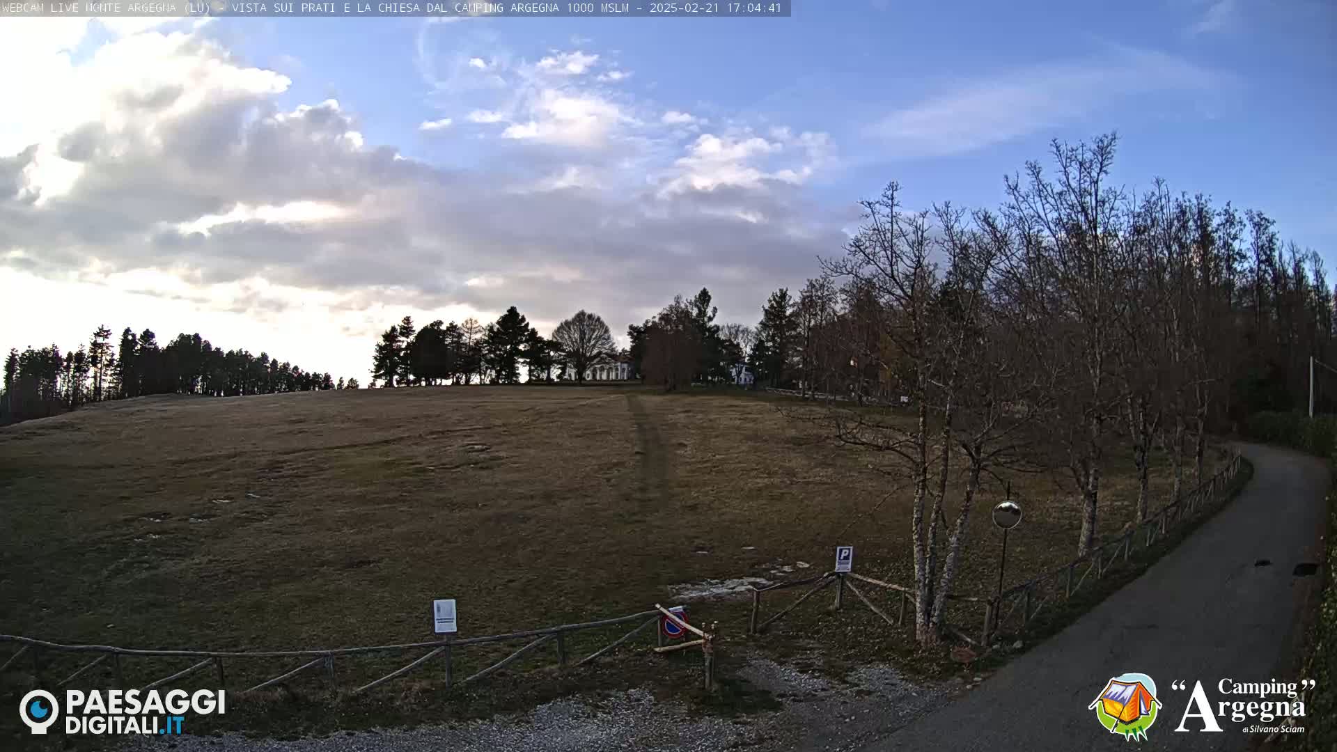 A mostly cloudy sky overlooks a brown, grassy field bordered by leafless trees and a curving road.