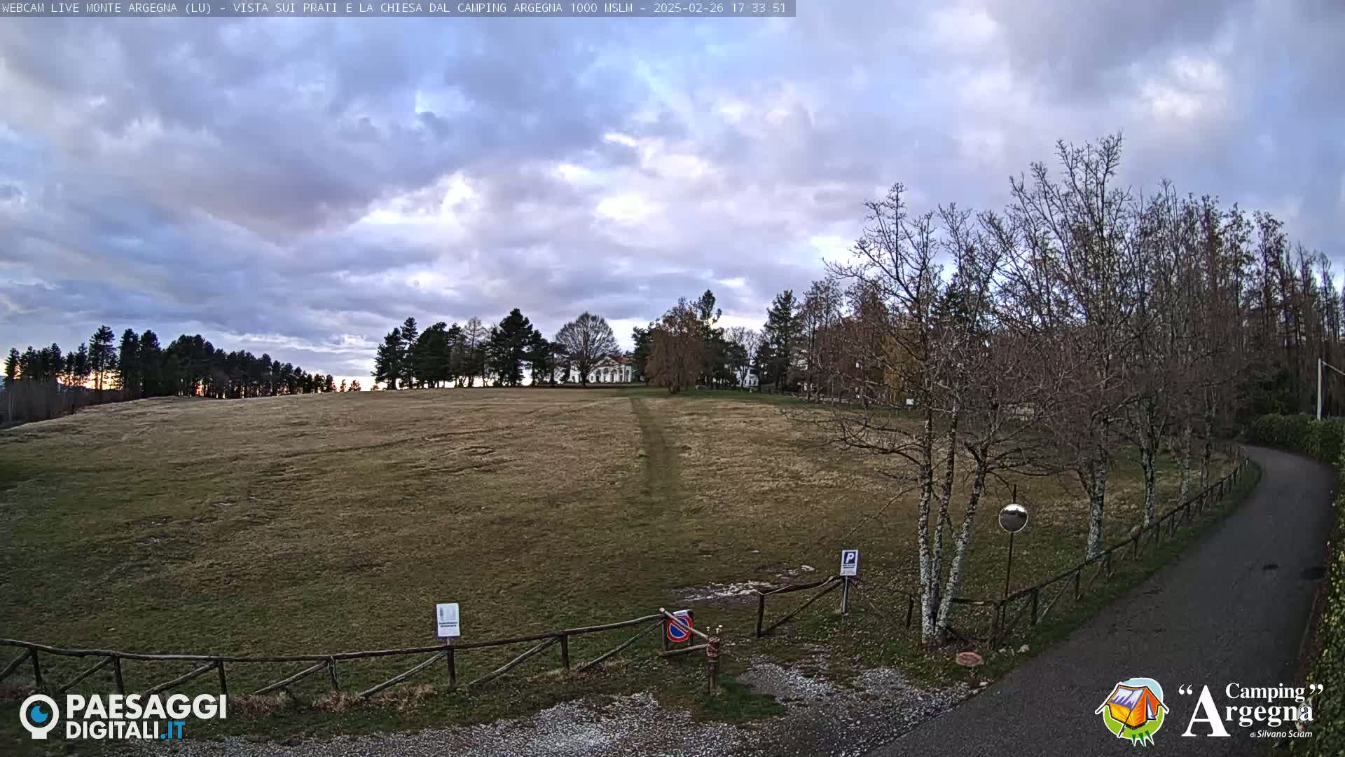 A mostly cloudy sky hangs over a brown, grassy field bordered by leafless trees and a paved road.