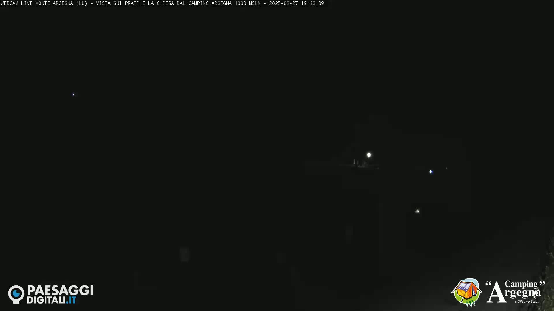 A nighttime, long shot of dimly lit, dark landscape with a few faint lights visible in the distance.